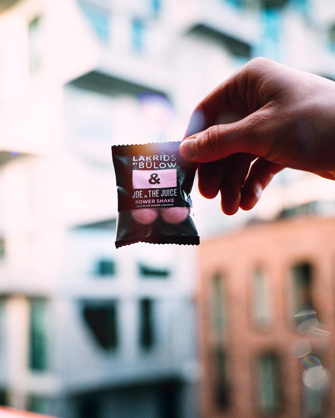 Close-up of a person's hand holding a small black snack package labeled 'Lakrids by Bülow & Joe & The Juice Power Shake' with a blurred urban background.