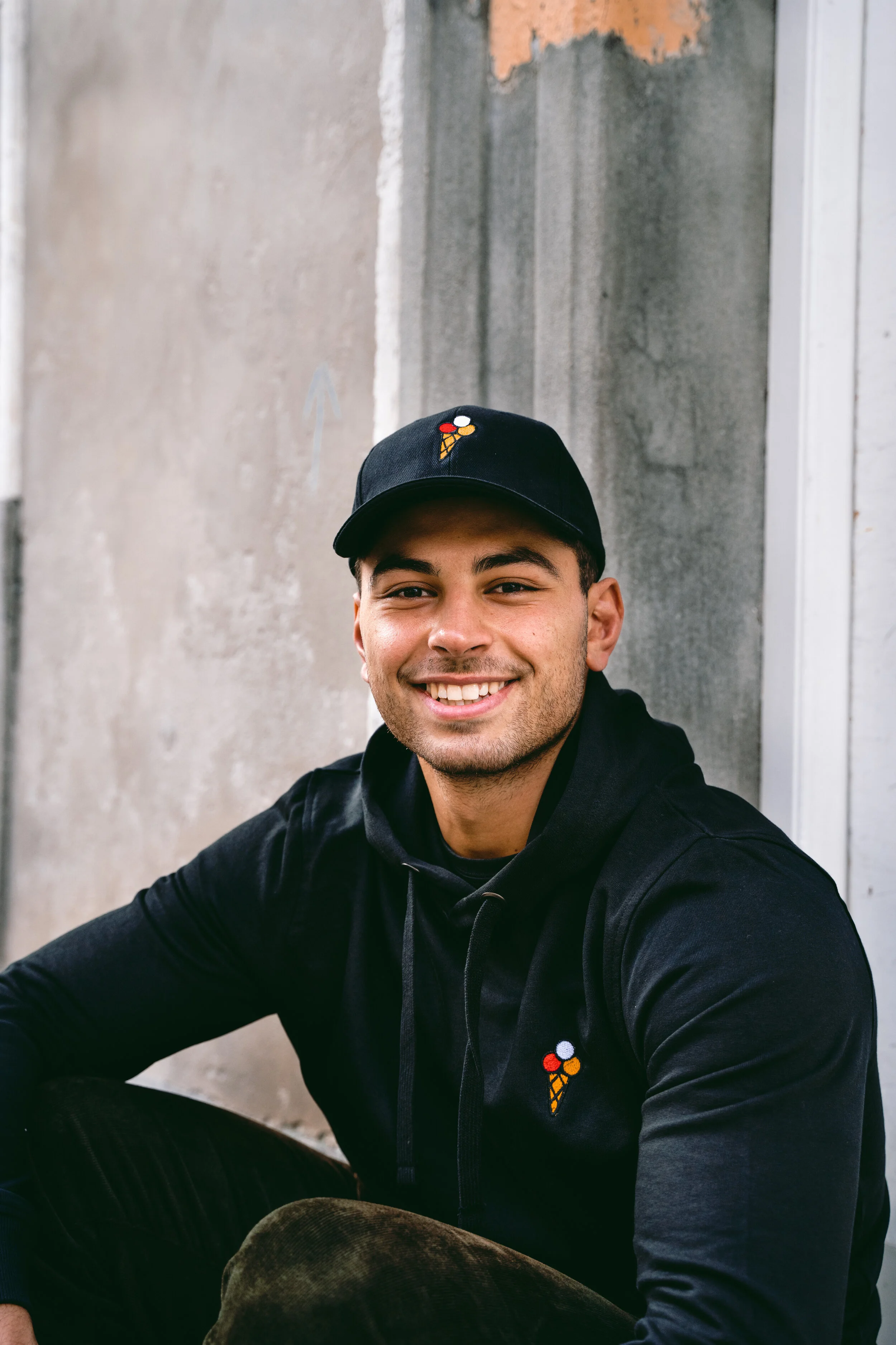 A young man with a smile, wearing a black cap and hoodie, sitting outdoors against a textured concrete wall.