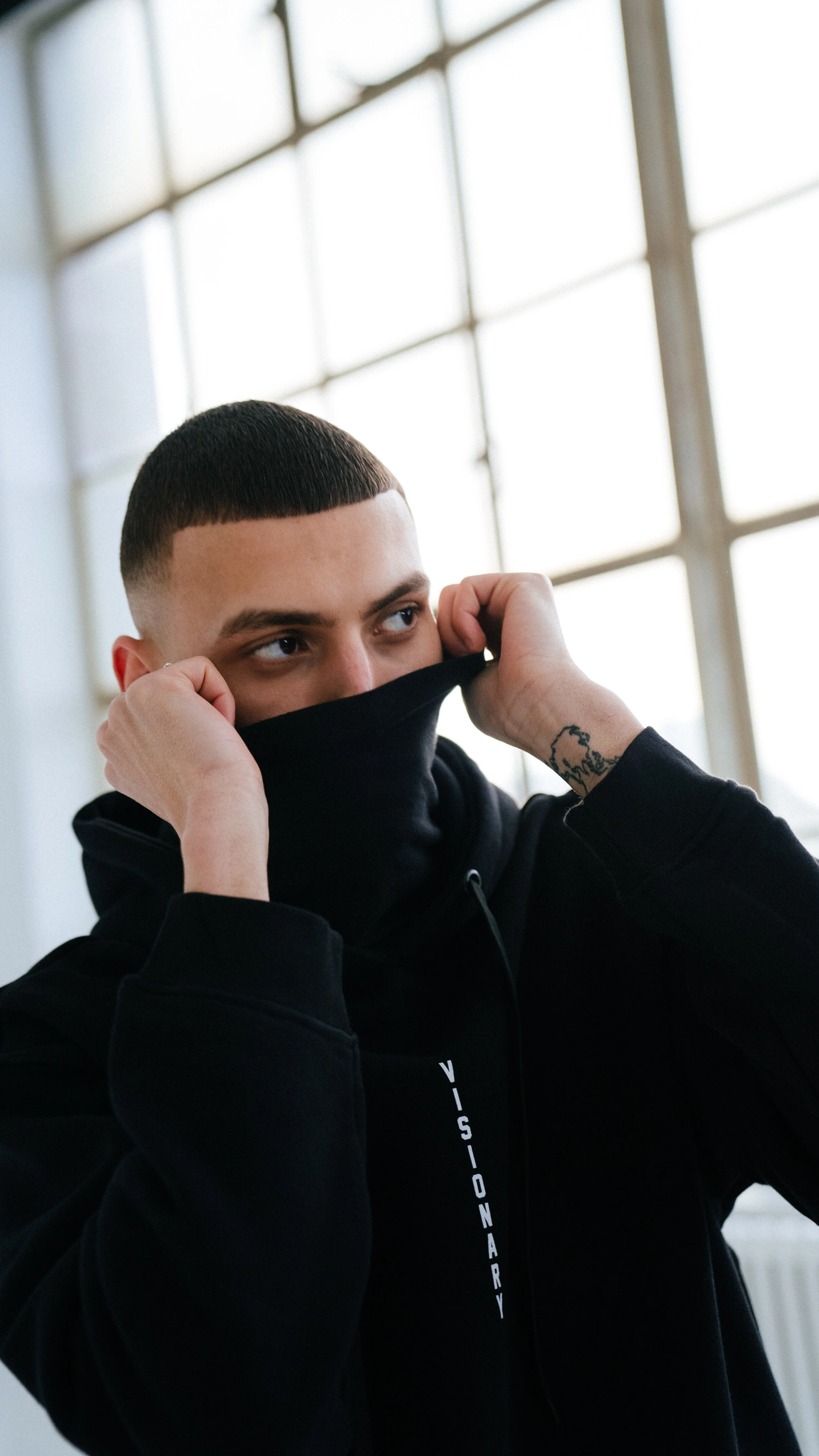 Young man with a buzz cut hairstyle and tattoos adjusts a black face mask with a hoodie that says 'VISIONARY'.