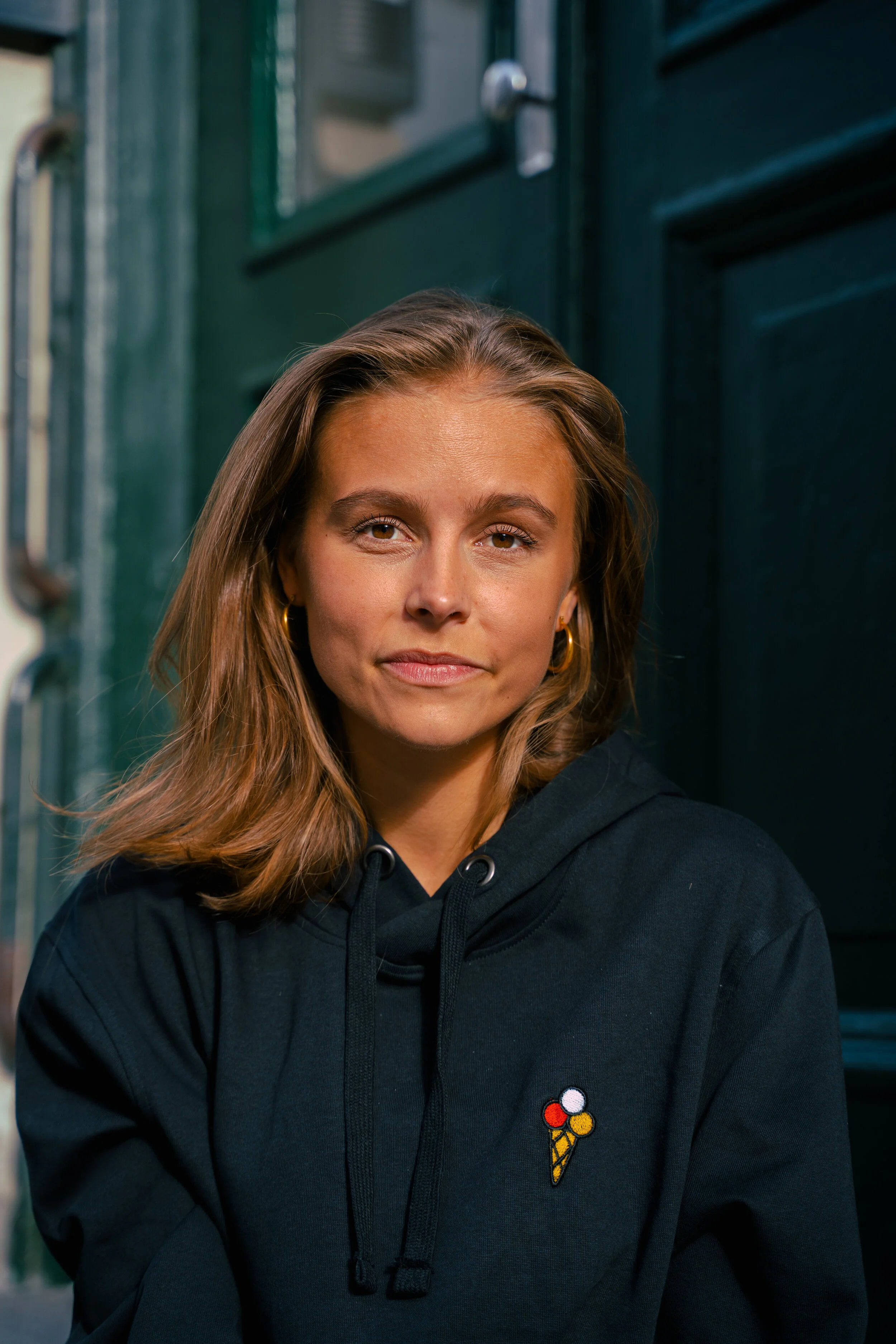 A young woman with light brown hair wearing a black hoodie with an ice cream cone embroidered on it, standing in front of a dark green door.