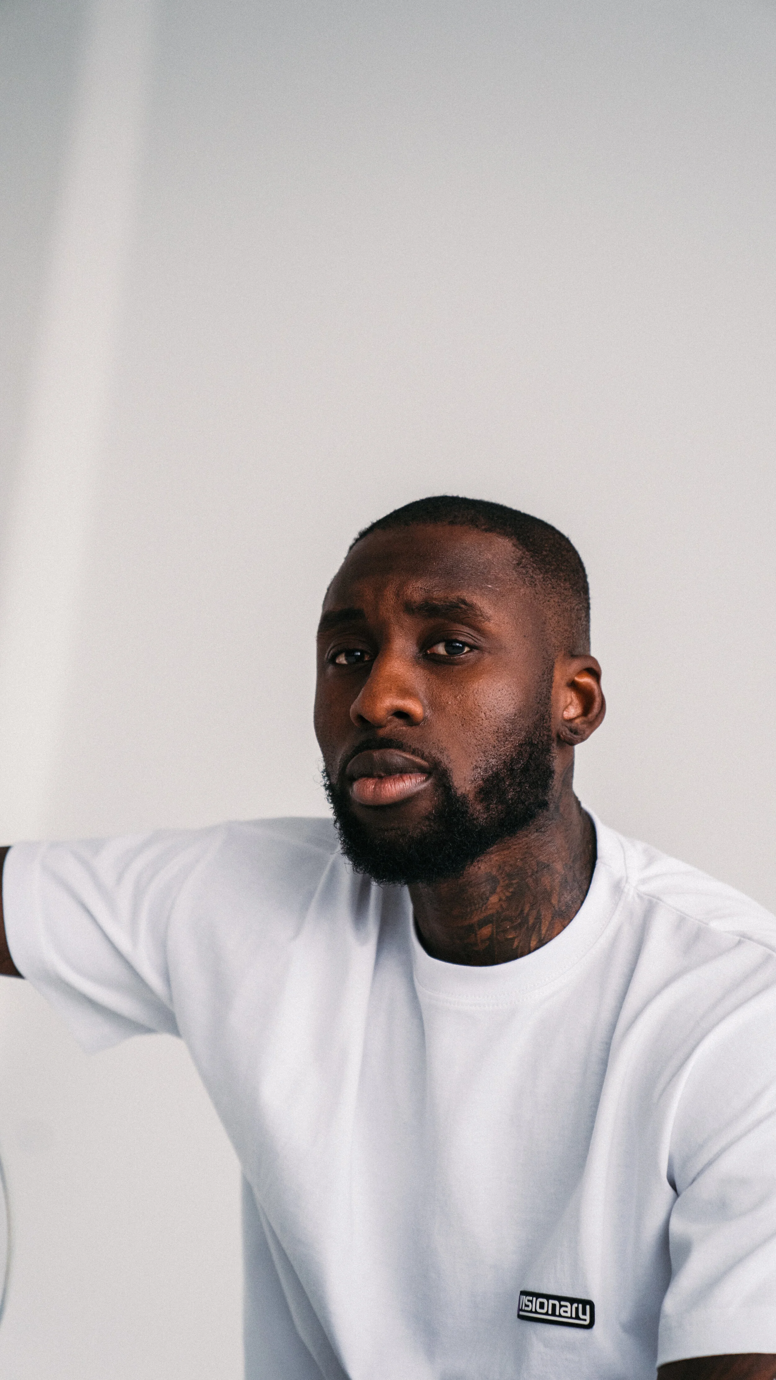 Portrait of a young man with short hair and beard, wearing a white t-shirt with the word 'VISIONARY' on it, against a plain light background.