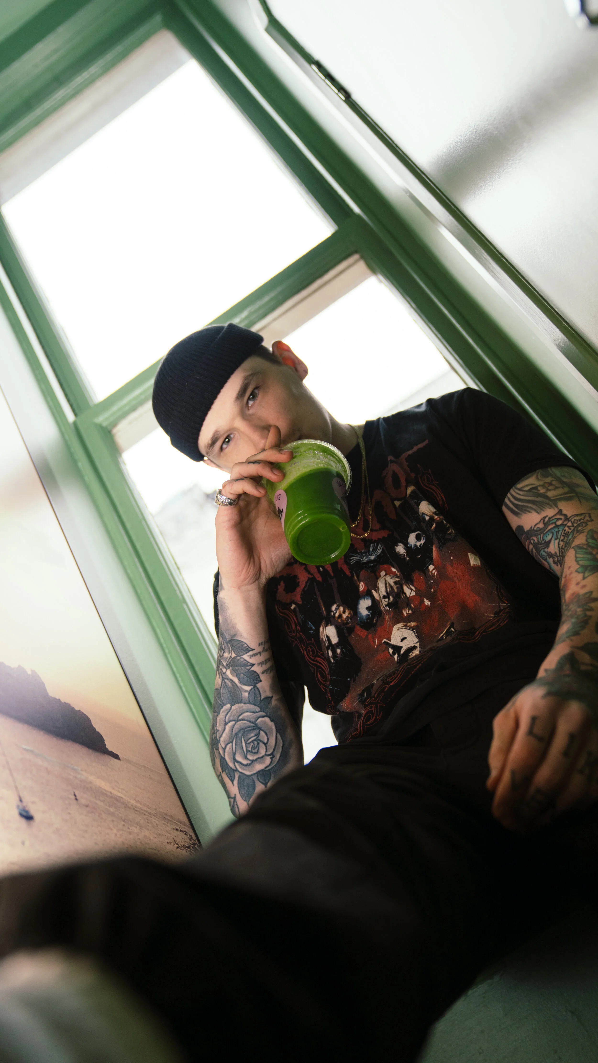 A young man with tattoos on his arms, wearing a black beanie and a graphic t-shirt, is sitting near a window and drinking a green smoothie.