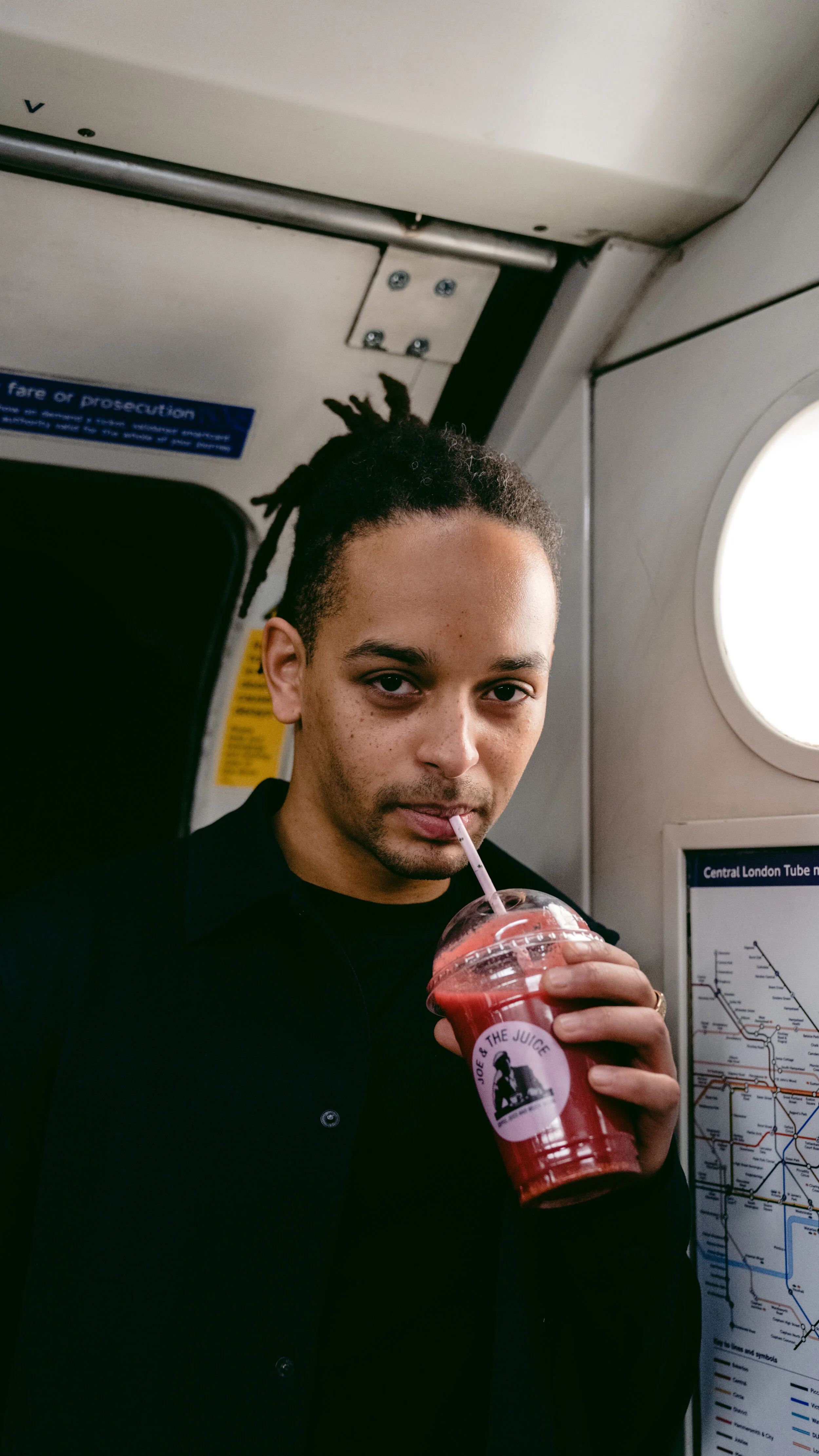 A man with dreadlocks drinking a red fruit smoothie through a straw on a subway train.