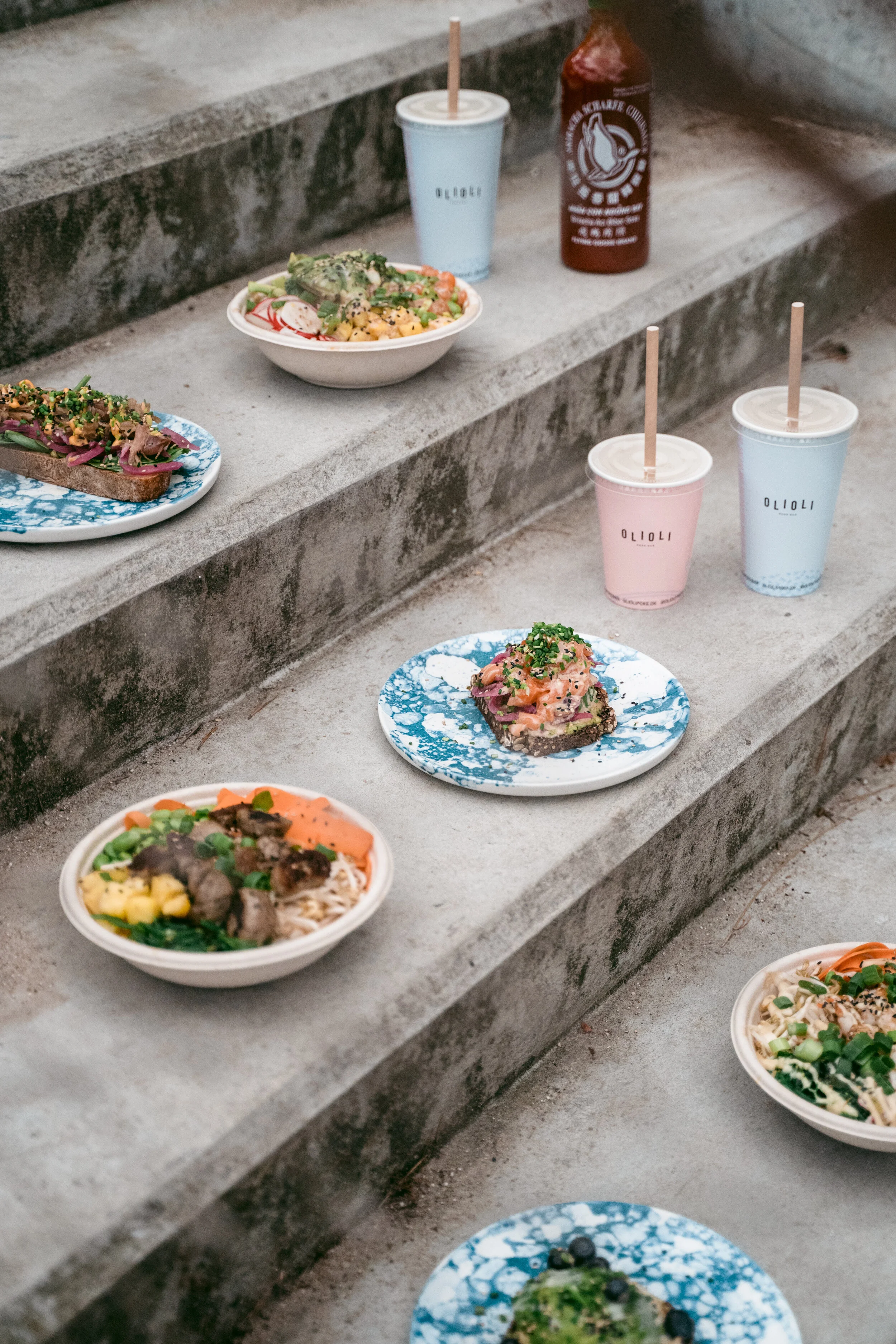 Food and drinks arranged on concrete stairs, including bowls of Asian dishes, toast with toppings, colorful beverages in cups, and a bottle of hot sauce.