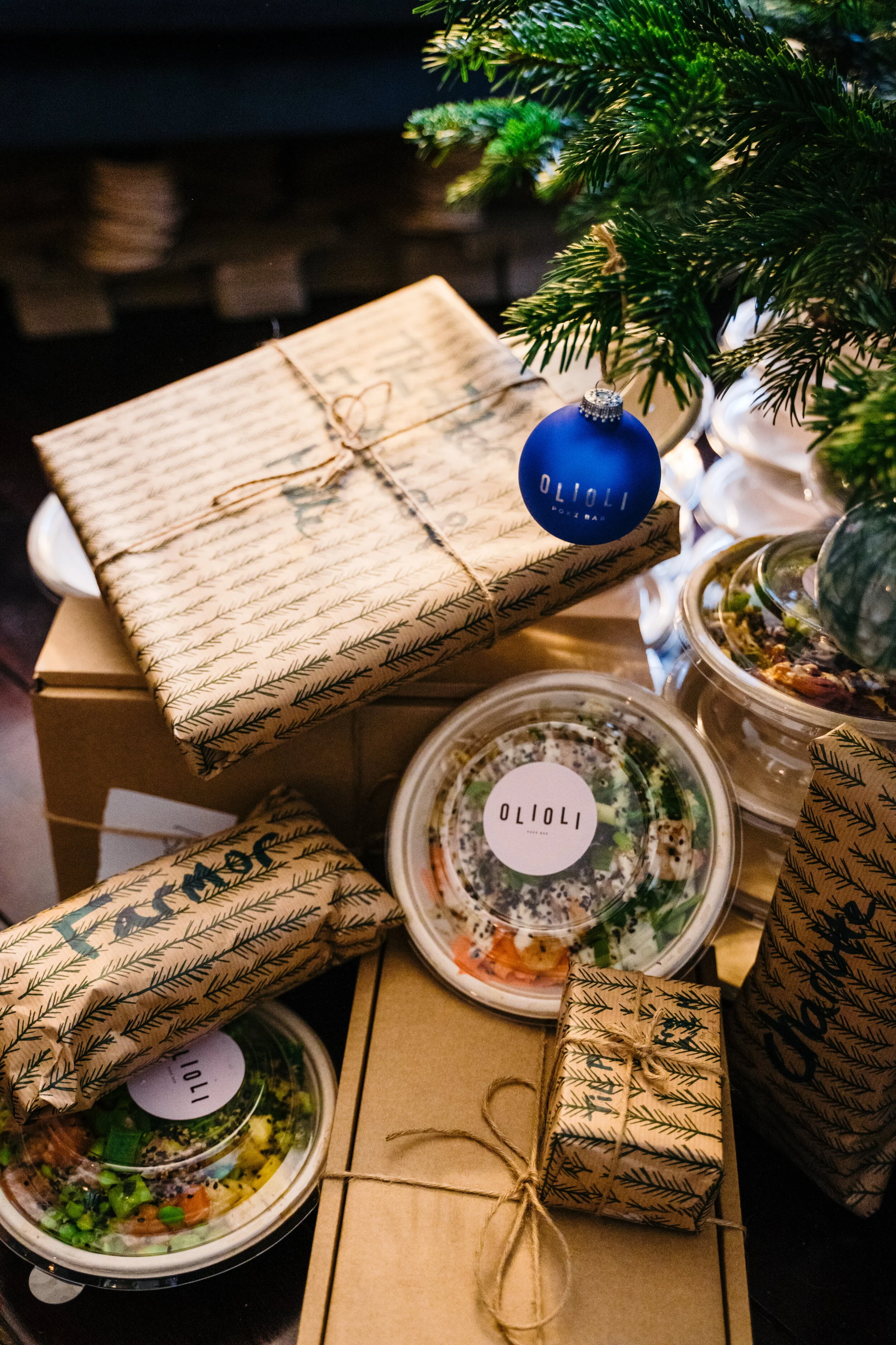 Holiday gifts and food containers under a green Christmas tree, with a blue ornament hanging from the tree