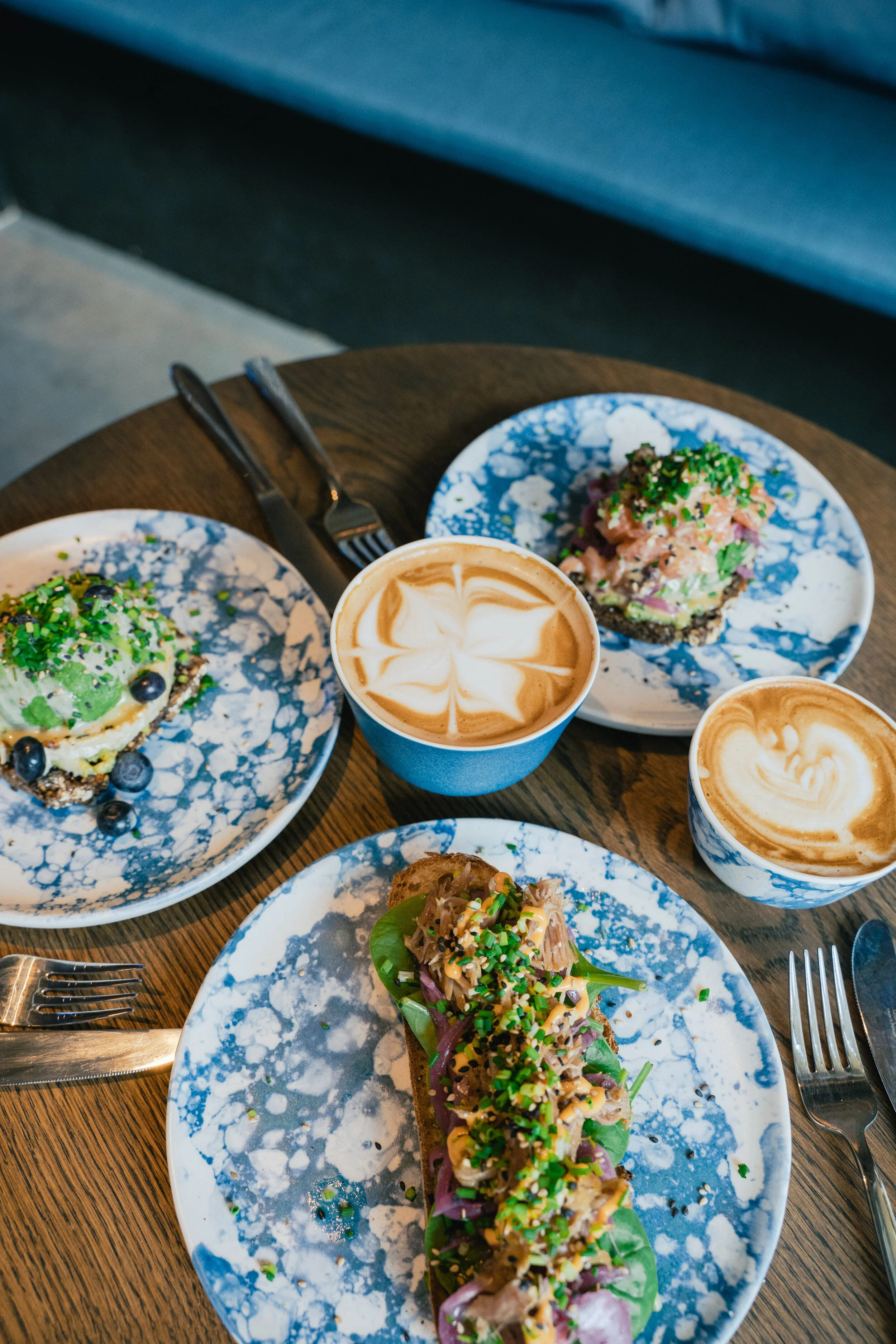Table with two plates of avocado toast topped with chopped herbs and seeds, two cups of coffee with latte art, and a cup of cappuccino in a cozy cafe setting.