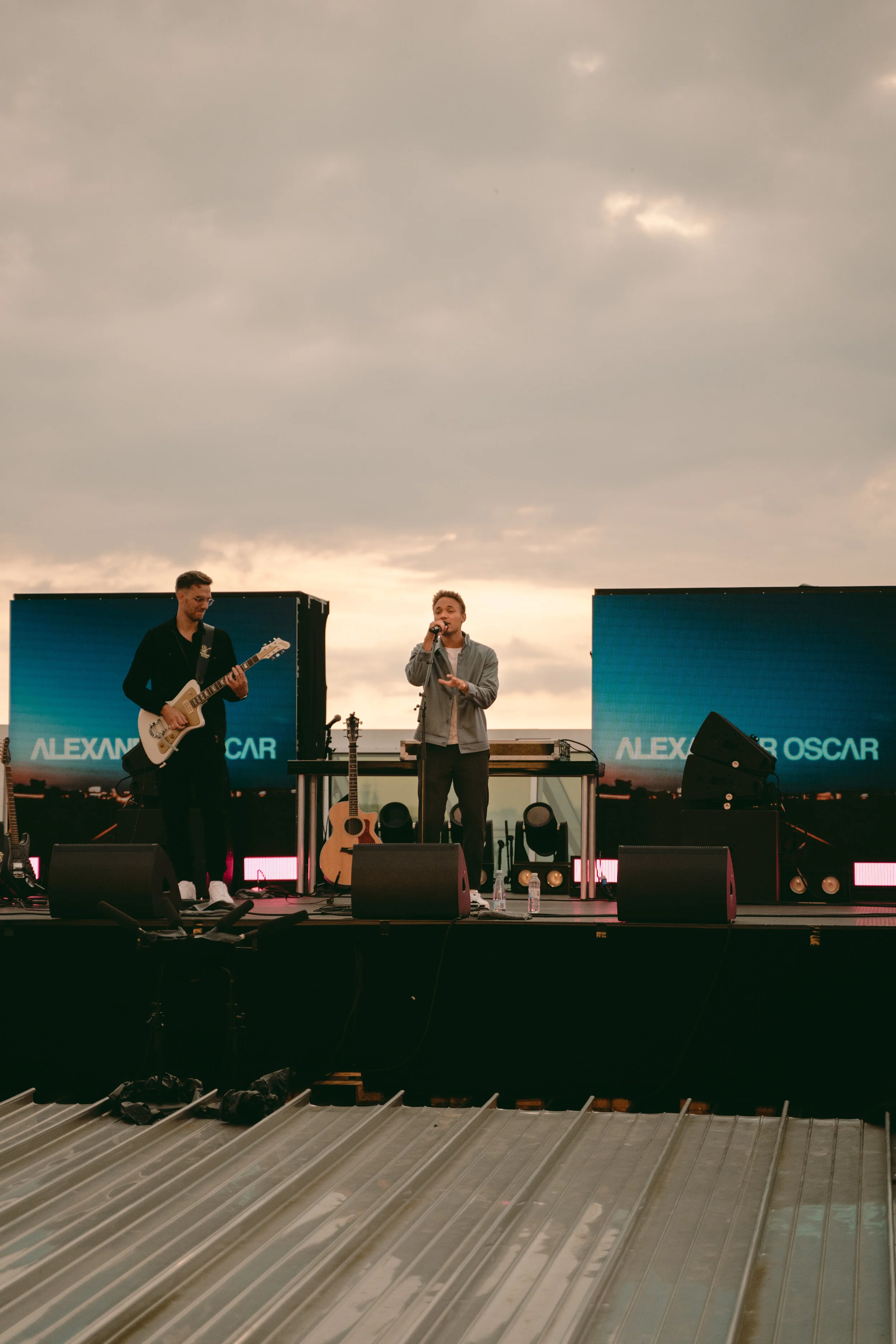 Two musicians performing on an outdoor stage at sunset, one playing guitar and the other singing into a microphone, with large screens behind them displaying the name 'Alexanio Oscar'.