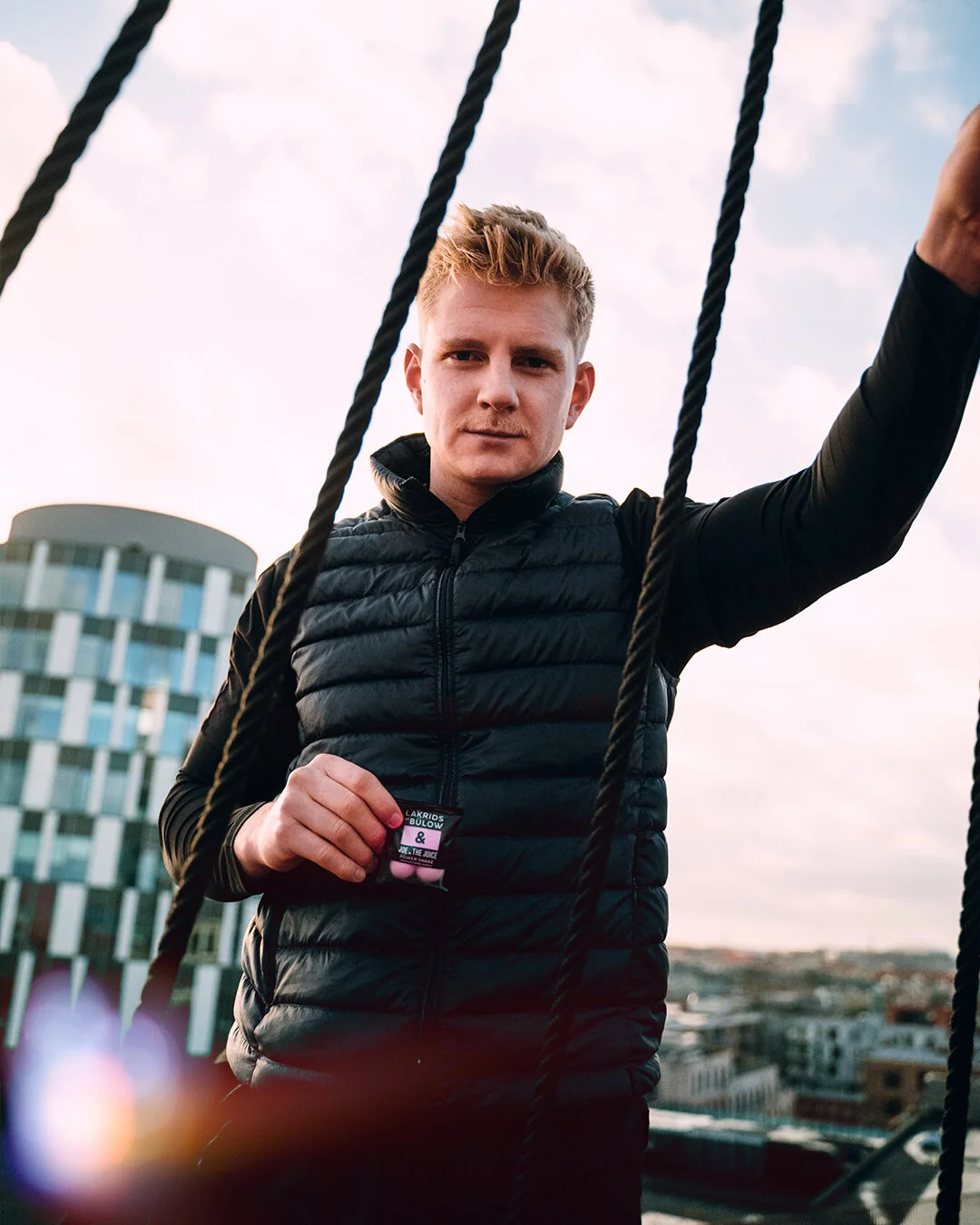 A young man in a black puffer vest standing on a rooftop, holding a small object, with city buildings in the background and a cloudy sky.