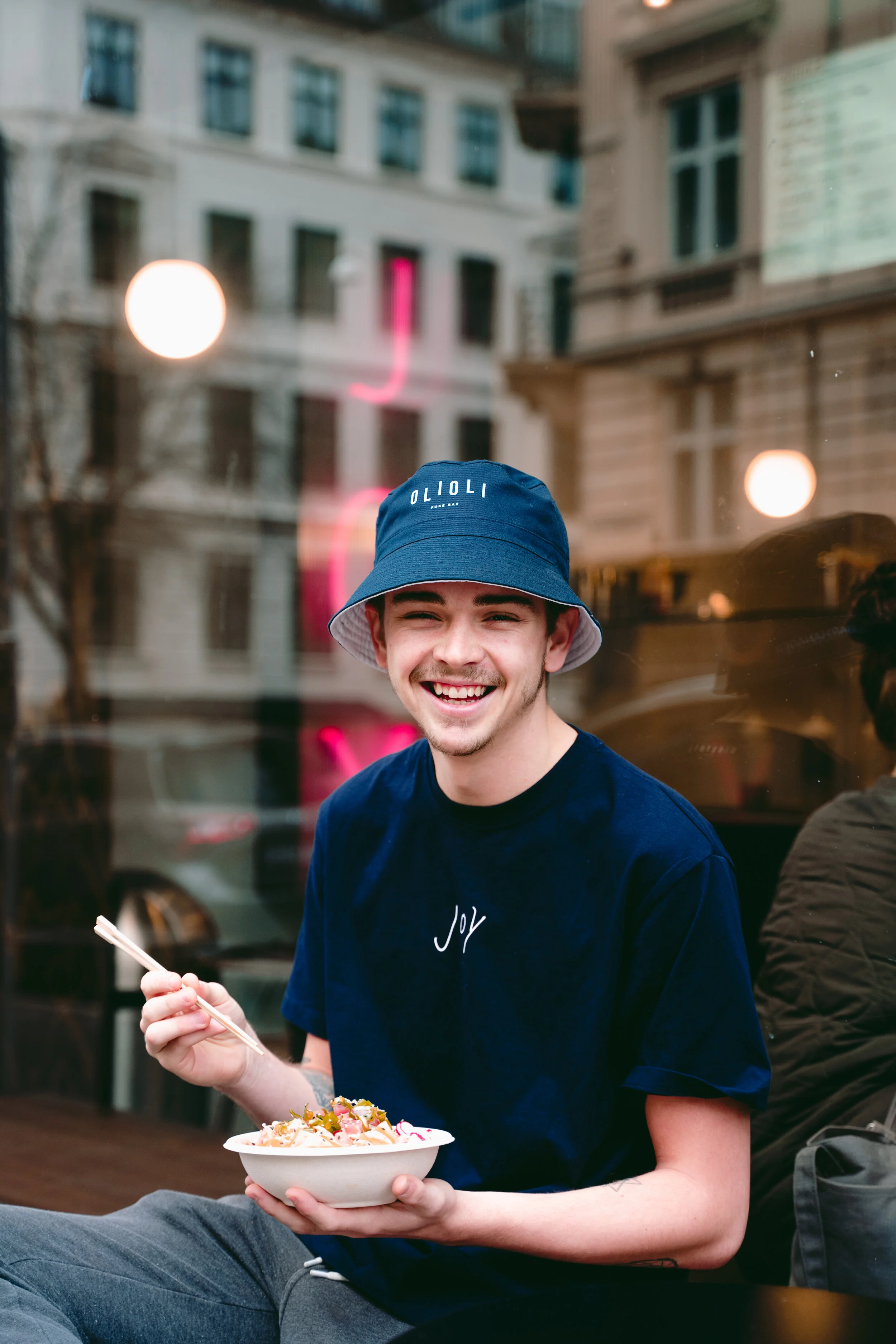 A young man is sitting outside a restaurant, smiling, and holding a bowl of food with chopsticks. He is wearing a blue bucket hat and a blue t-shirt with the word 'Joy' printed on it. The background has a glass window reflecting the street scene.