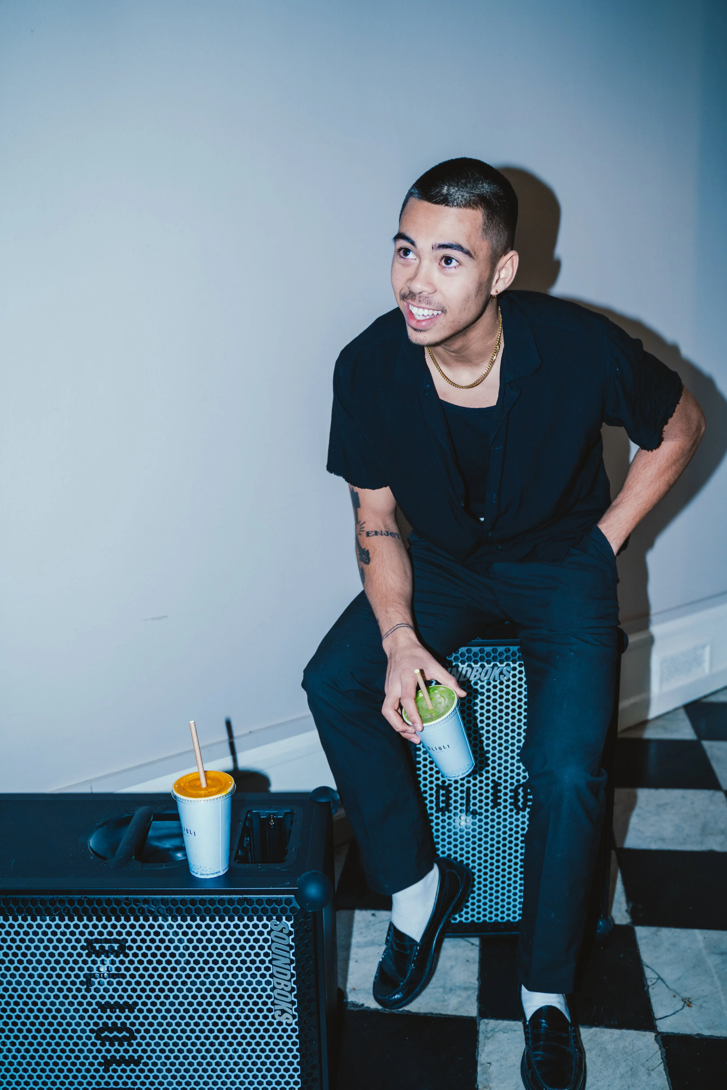 A young man with short hair, dressed in black, sitting on a speaker, smiling and holding a drink. There is another drink on the speaker next to him.