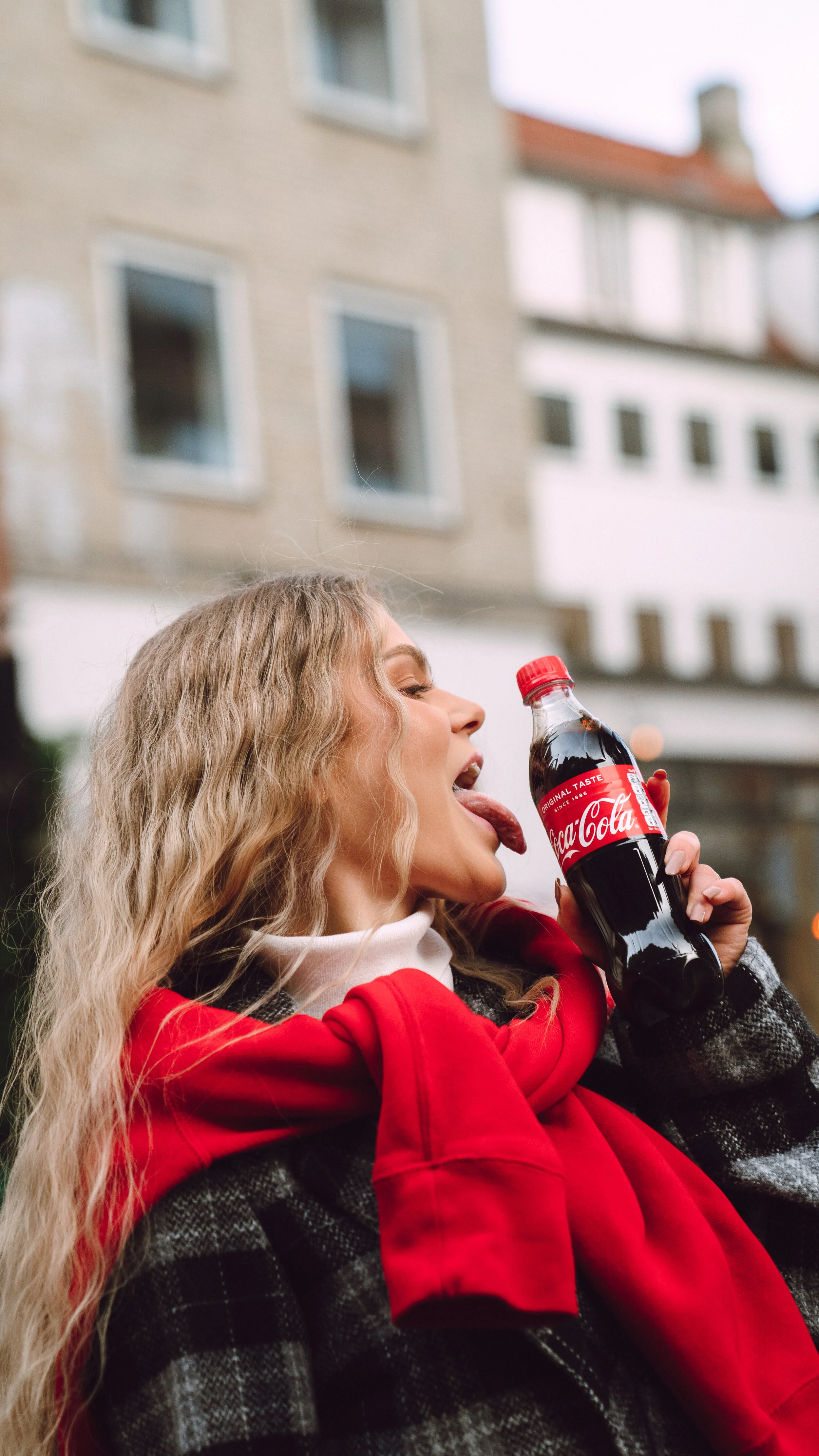 Young woman with long curly blonde hair wearing a red hoodie and black checkered jacket, holding a bottle of Coca-Cola, licking it with her tongue, standing outdoors with buildings in the background.