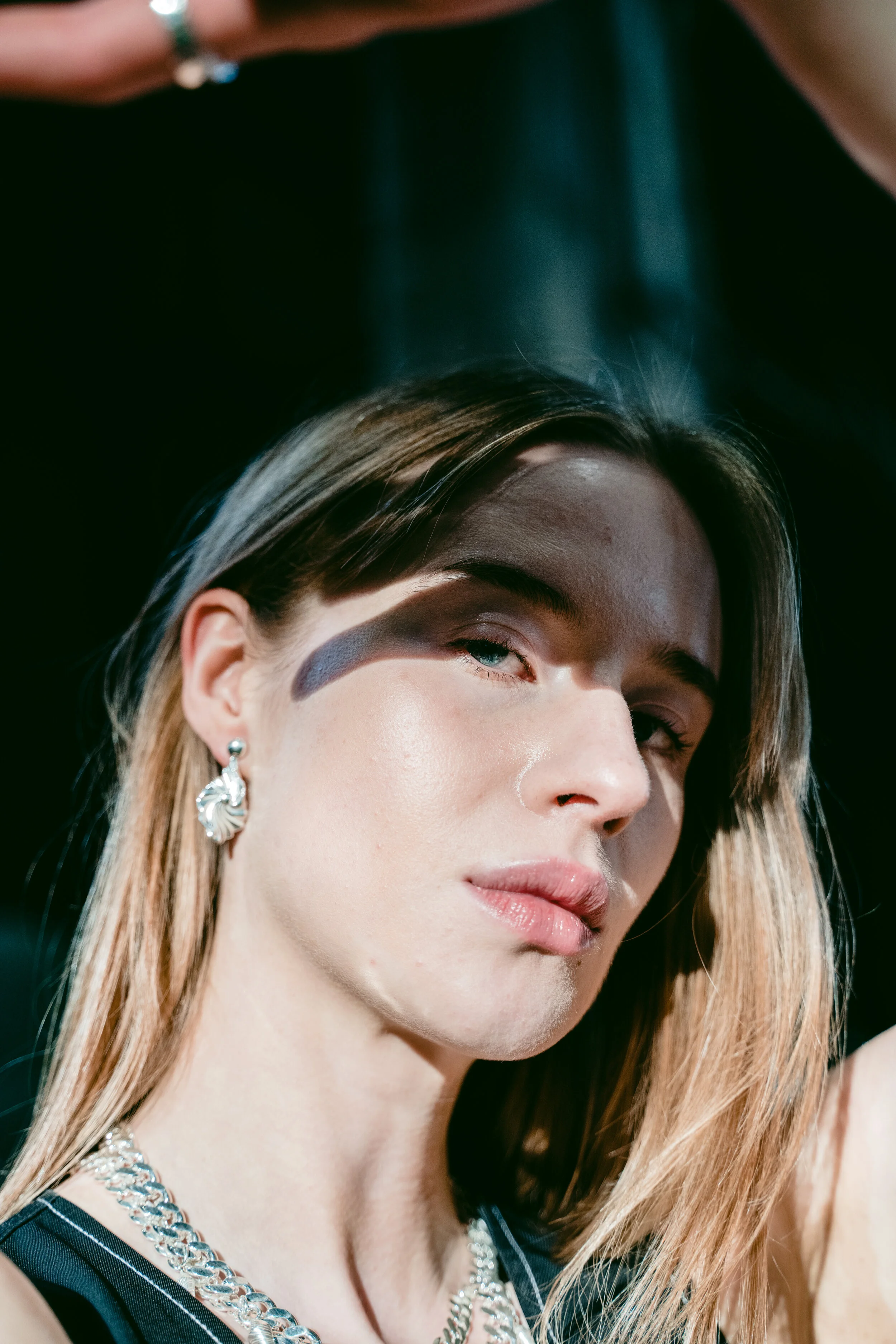 Close-up of a young woman with light skin and blonde hair, wearing silver jewelry, with a partial dark background.