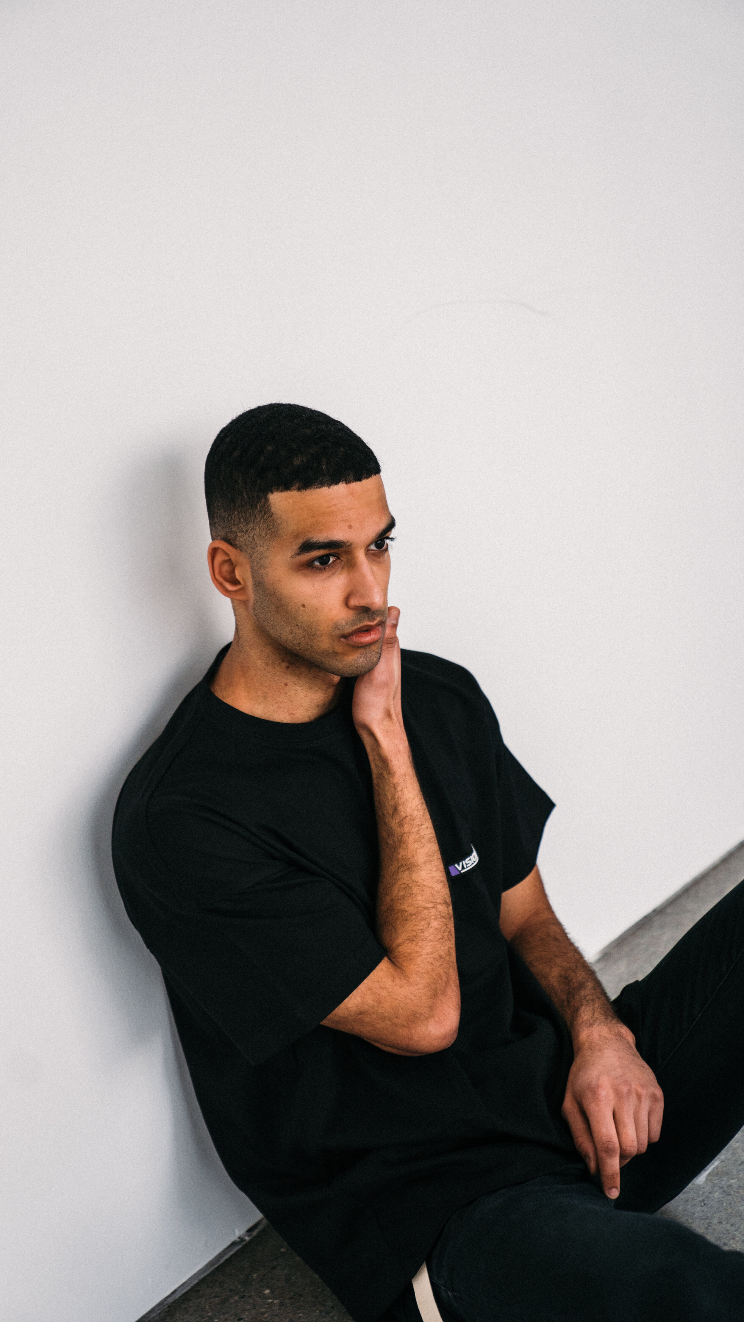 A young man sitting on the floor, leaning against a plain white wall, with one hand on his neck and a thoughtful expression.