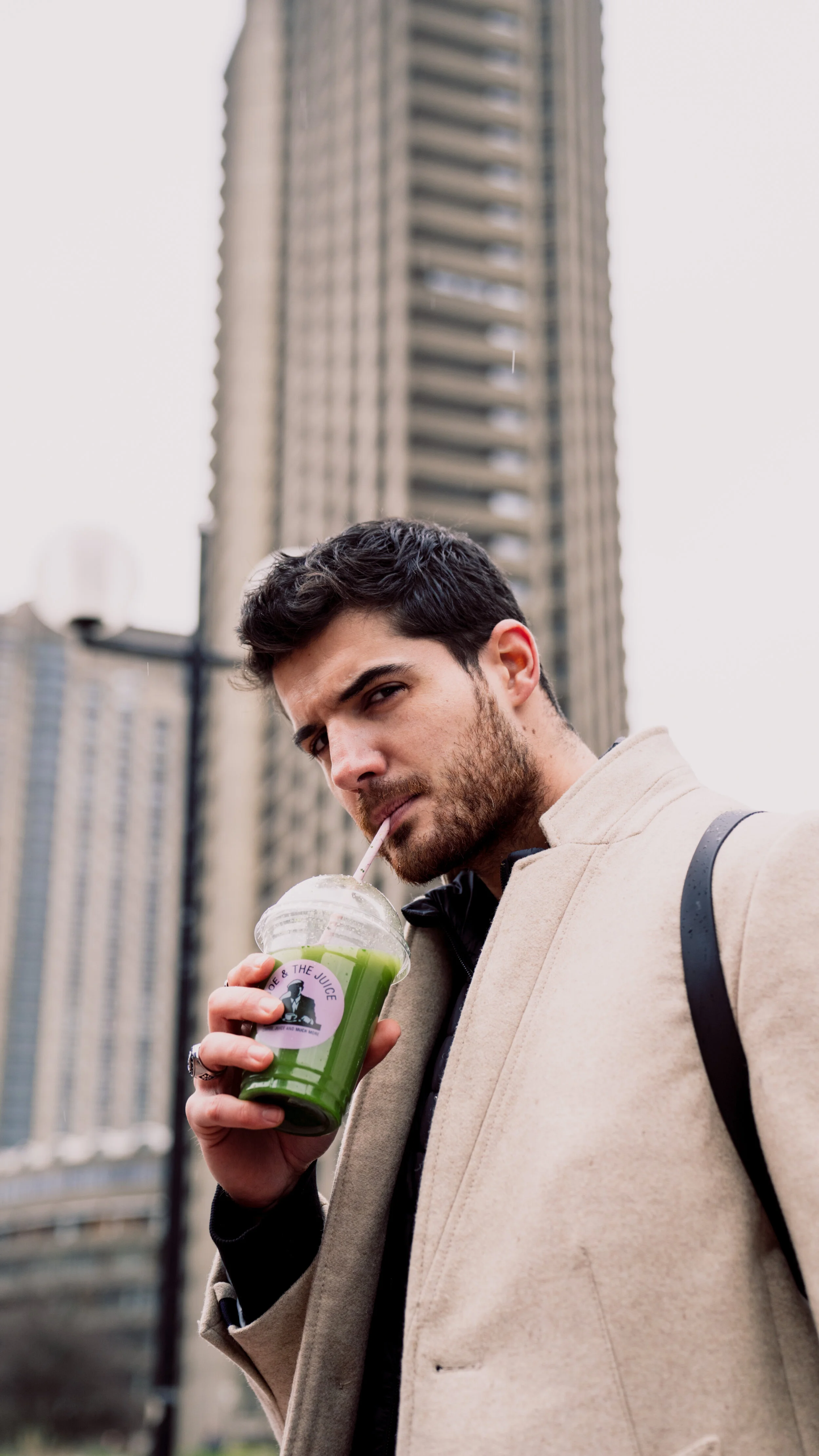 A young man with dark hair and a beard drinks a green smoothie from a clear plastic cup with a straw, standing outdoors in front of tall buildings on a city street.