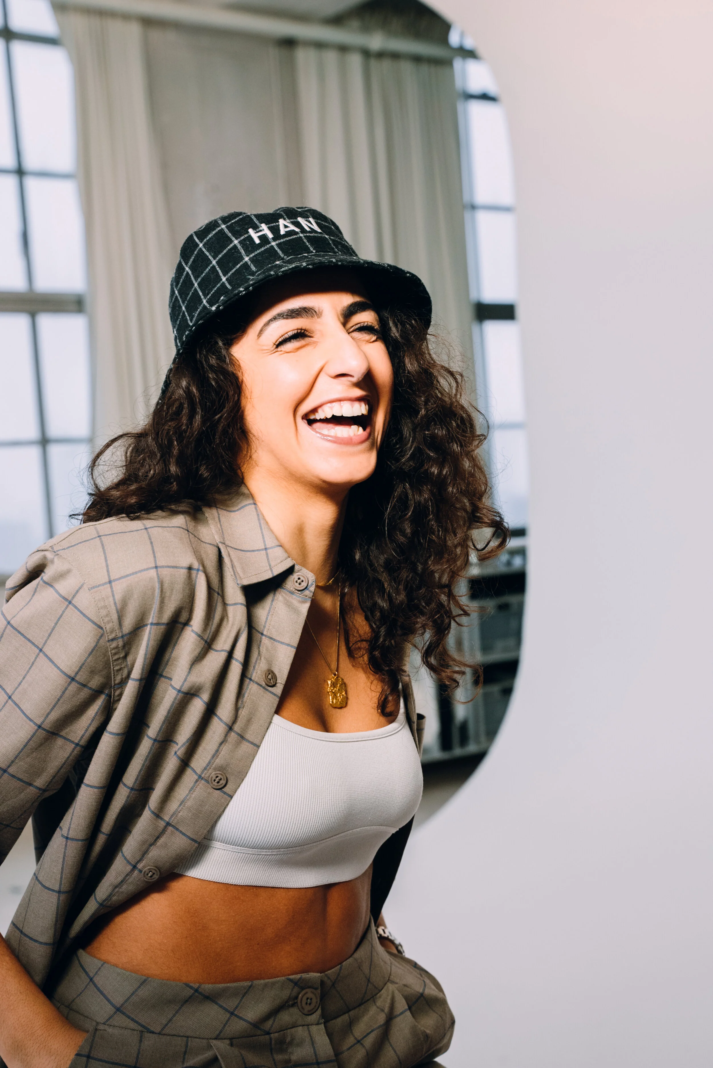 A woman with curly dark hair, smiling and laughing, wearing a plaid bucket hat with the word 'HANI' on it, a beige plaid blazer, a white crop top, and gold jewelry, in a modern indoor space with large windows and white walls.
