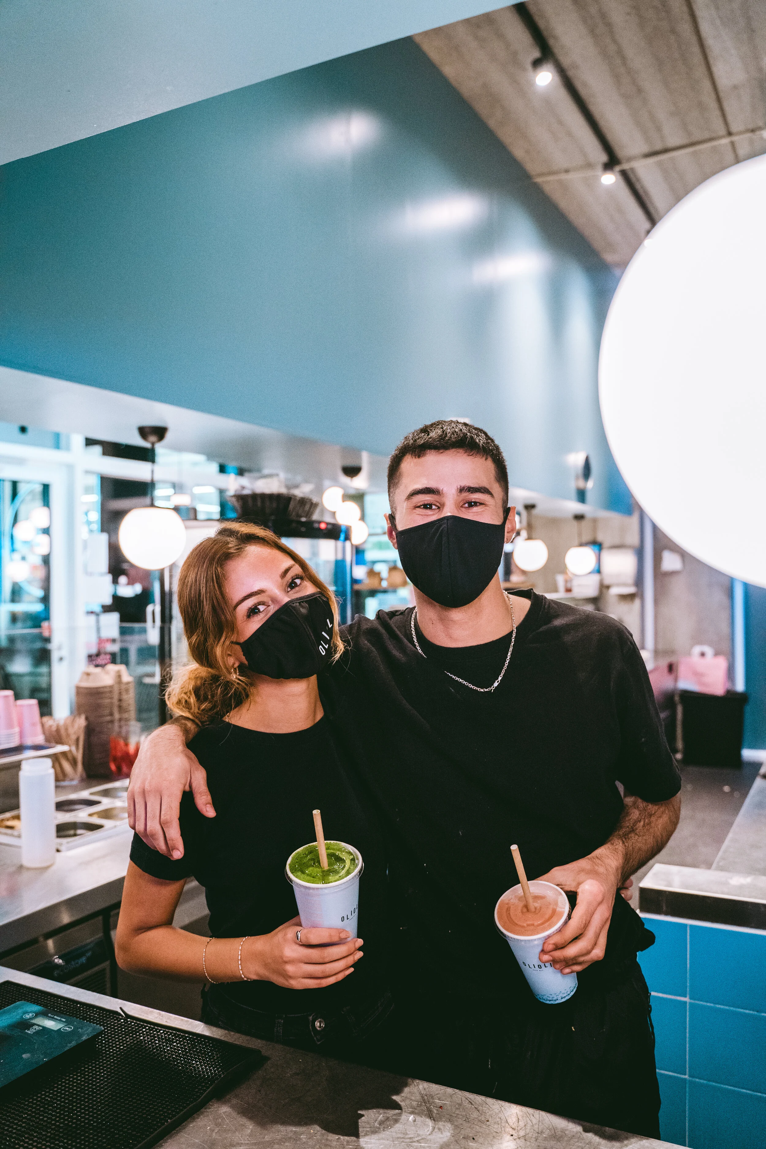 Two people wearing black masks and black shirts, smiling with one arm around each other, holding colorful drinks with straws, inside a modern cafe or juice bar.