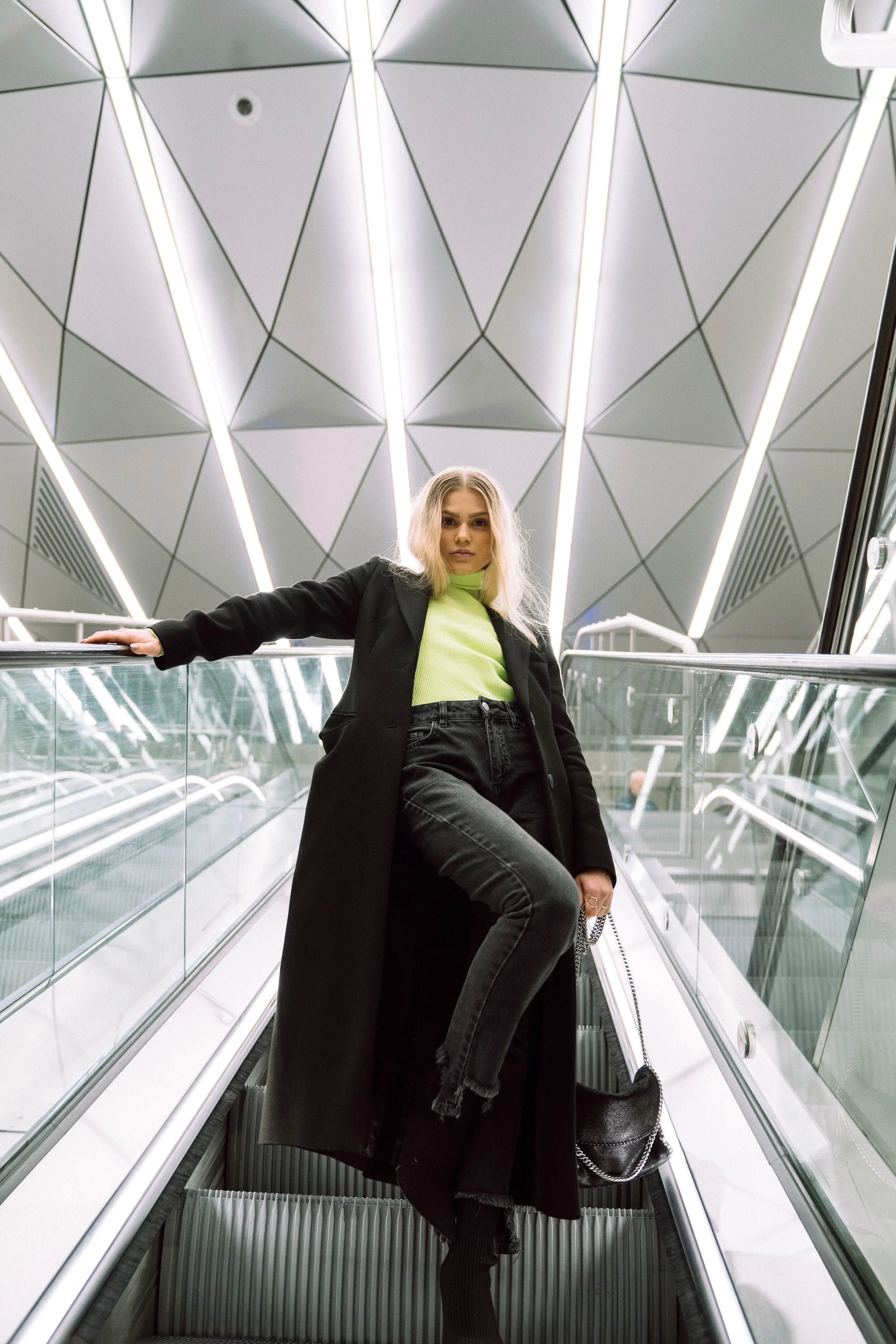 A young woman with blonde hair wearing a black coat and black jeans standing on an escalator in a modern building with geometric ceiling lights.