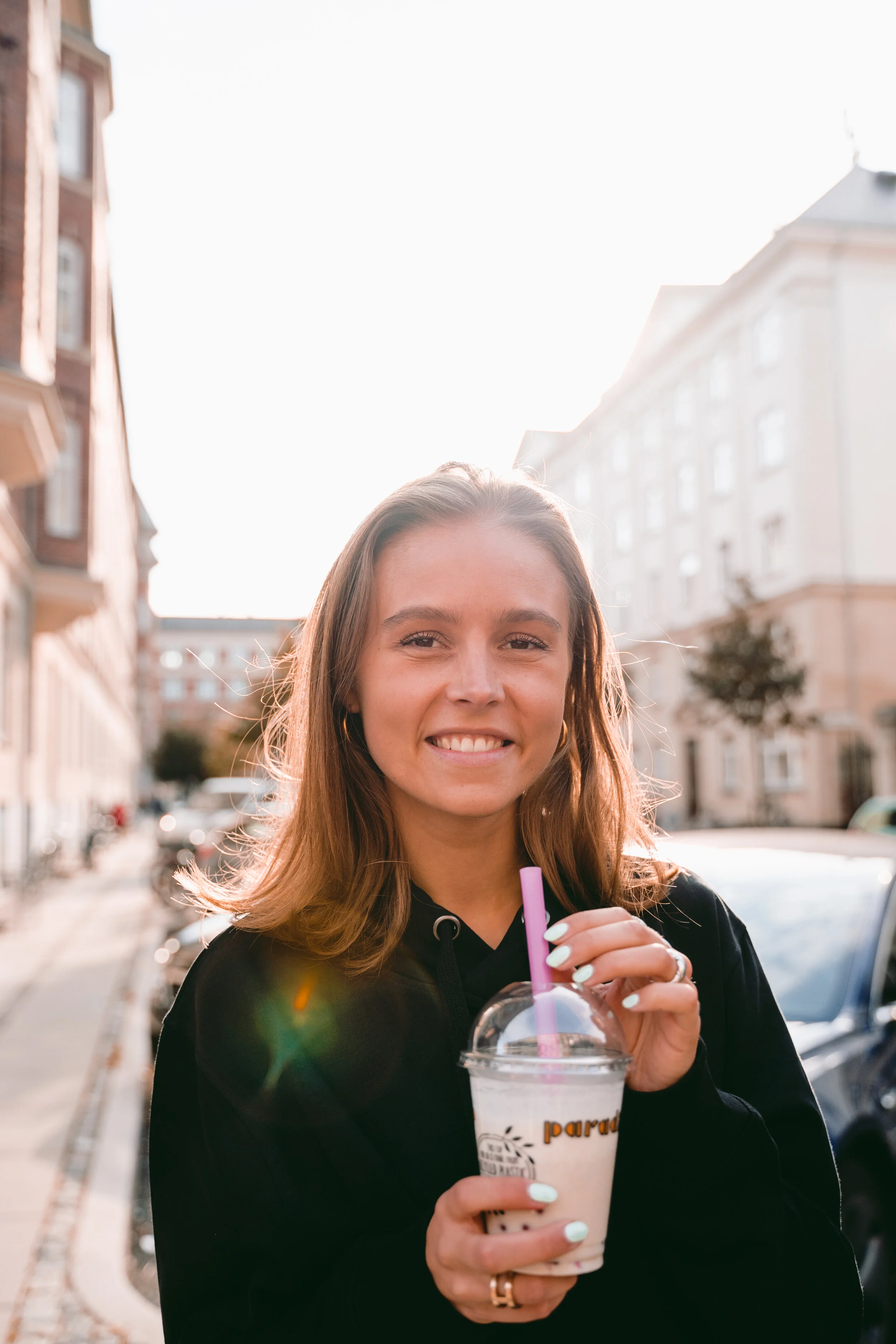Young woman with light brown hair smiling and holding a smoothie with a purple straw outdoors on a sunny day, with buildings and parked cars in the background.