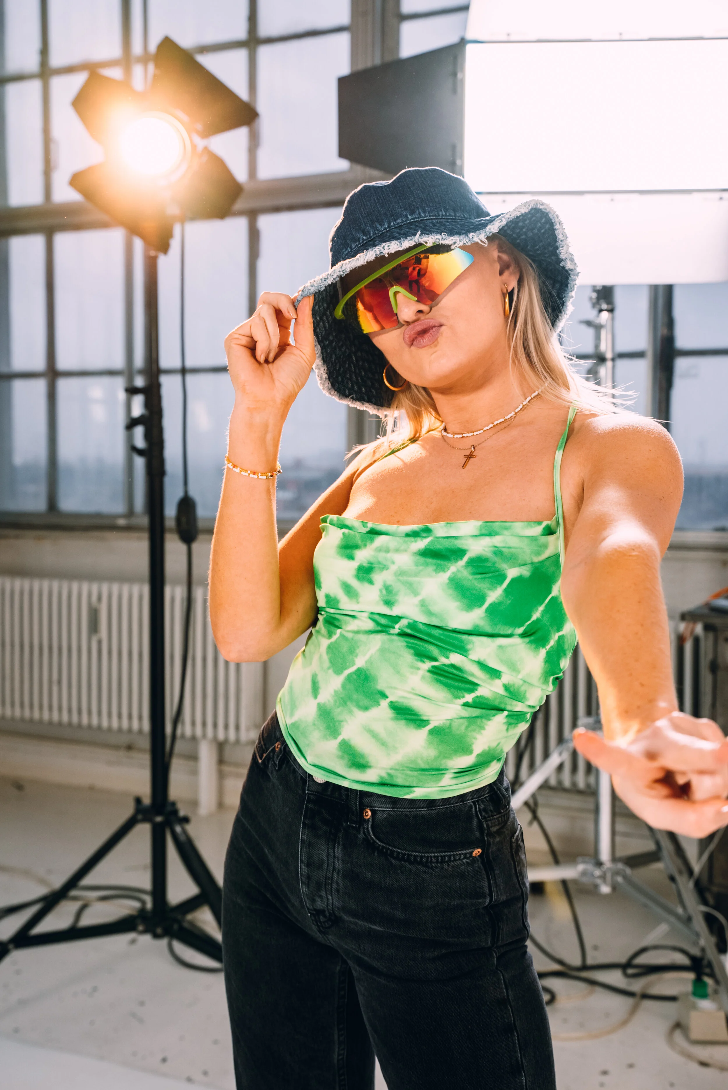 Woman in a green tie-dye tank top, black jeans, large sun hat, and reflective sunglasses in a photography studio with studio lights and equipment.