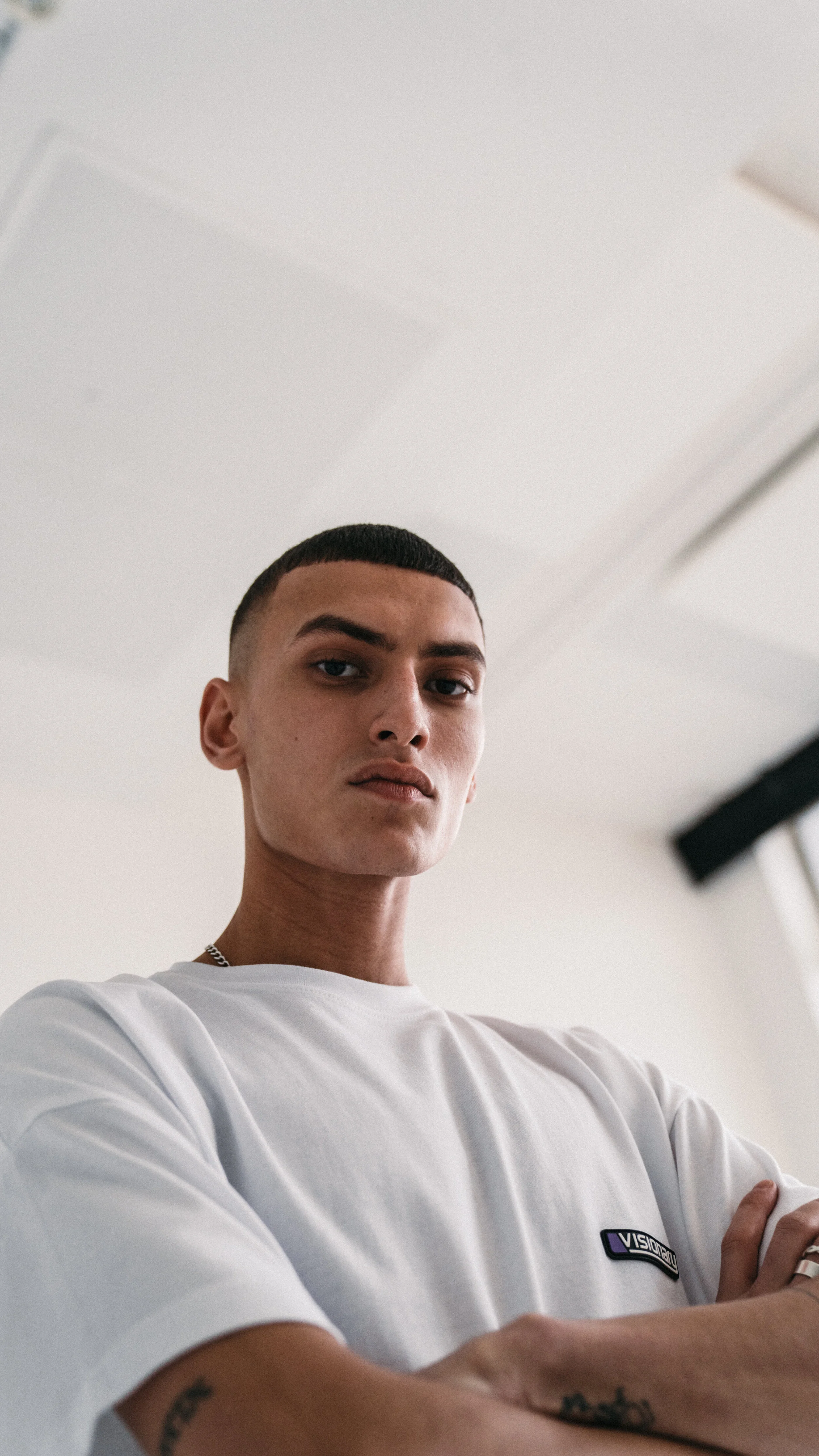 A young man with short dark hair, wearing a white t-shirt with a small black logo on the chest, standing indoors with arms crossed, looking directly at the camera.