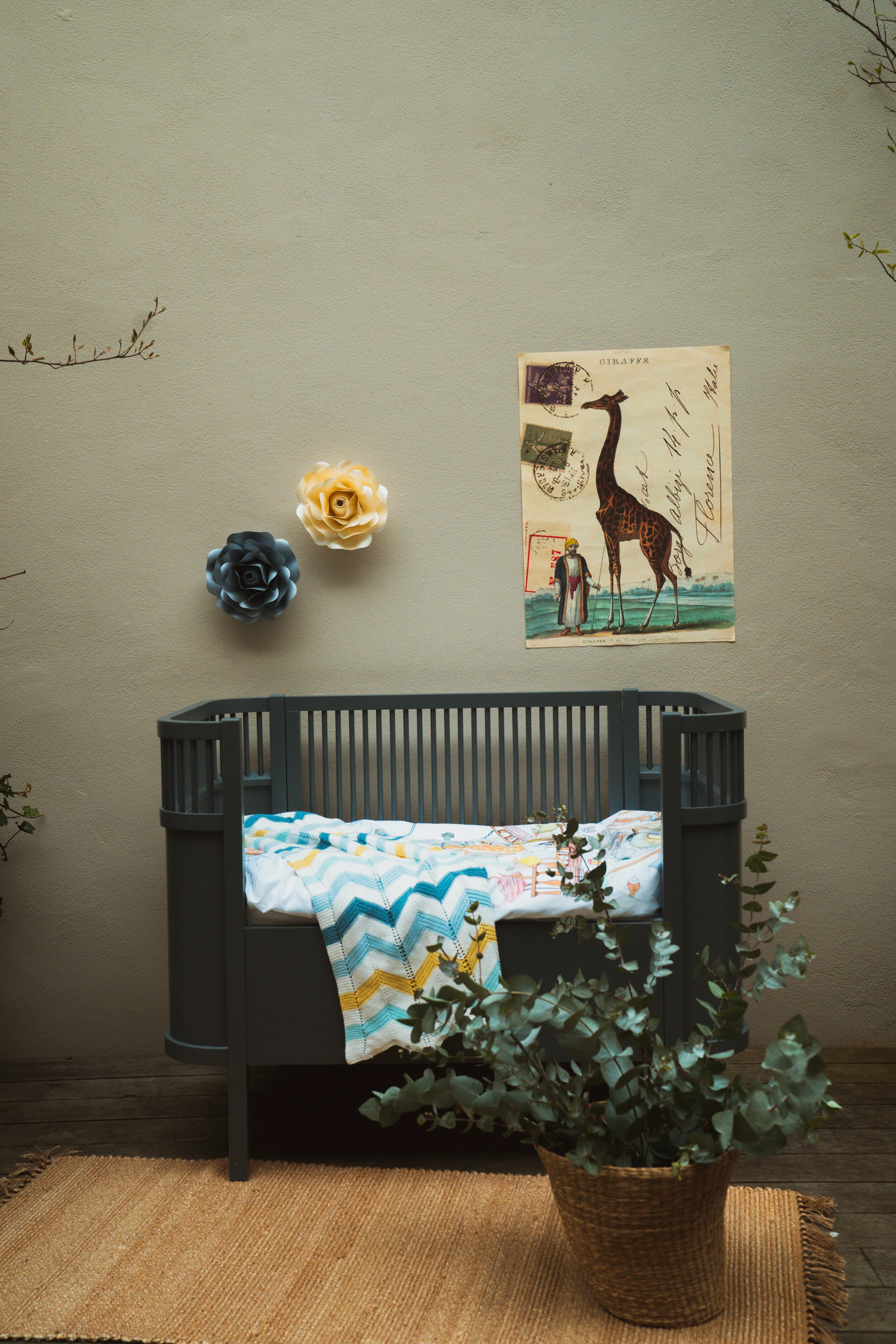 A nursery room with a gray crib, a colorful blanket, a potted eucalyptus plant, two paper flowers on the wall, and a vintage giraffe and person poster.