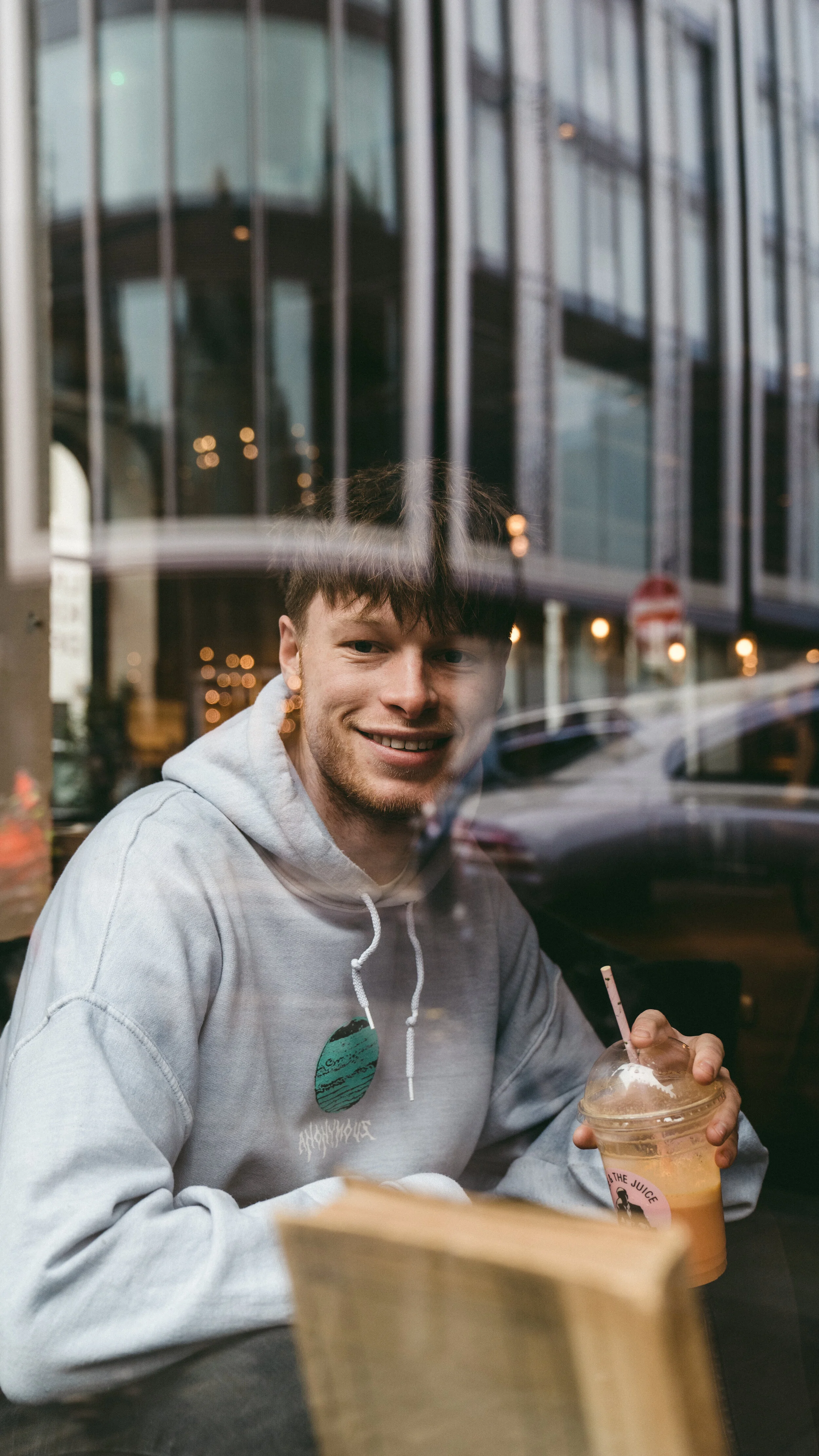 A young man with short brown hair and a gray hoodie, smiling and sitting inside a cafe, holding a plastic cup with iced tea and a straw, seen through the window reflection.