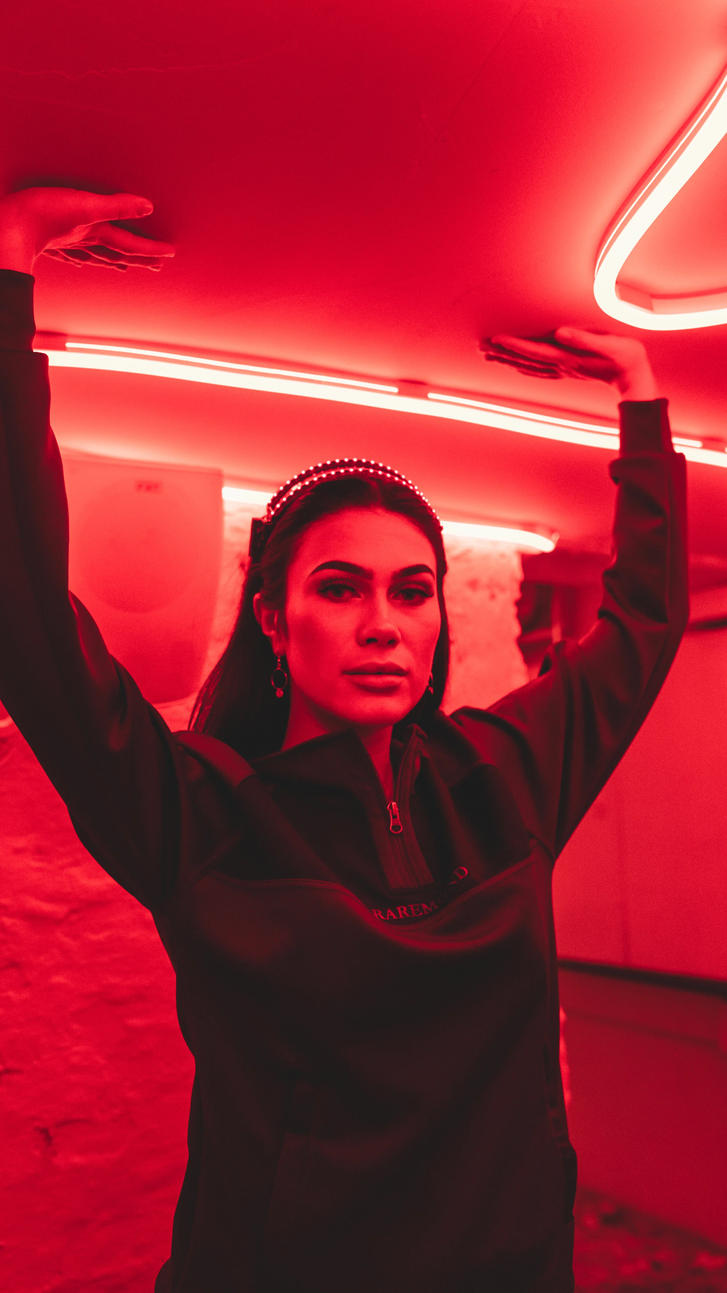 A woman standing in a red-lit room with her arms raised, wearing a black jacket with a zipper, earrings, and a headband.