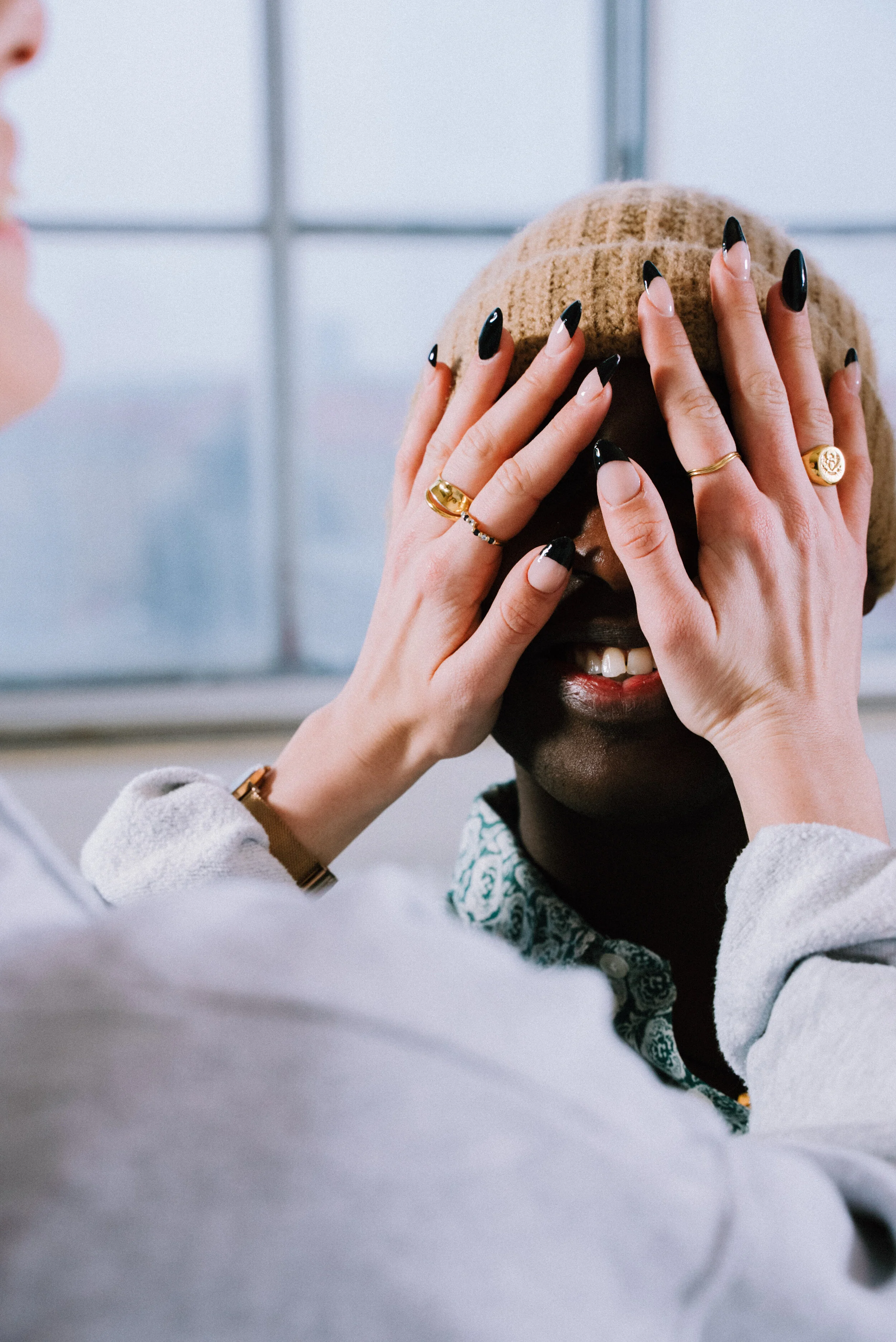 Two people playing peekaboo with a person wearing a beige knit hat, smiling, indoors near a window.