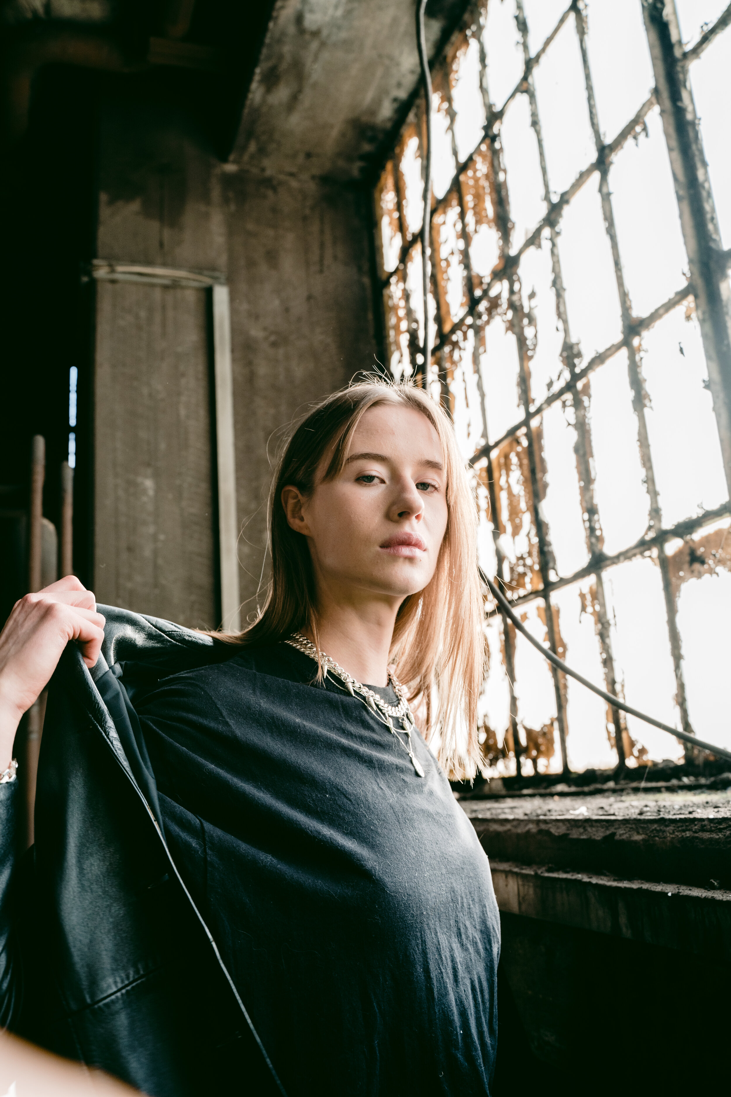 A young woman with blonde hair in a black t-shirt and layered necklaces, holding a leather jacket, stands inside a dilapidated building with burnt and broken orange window frames.