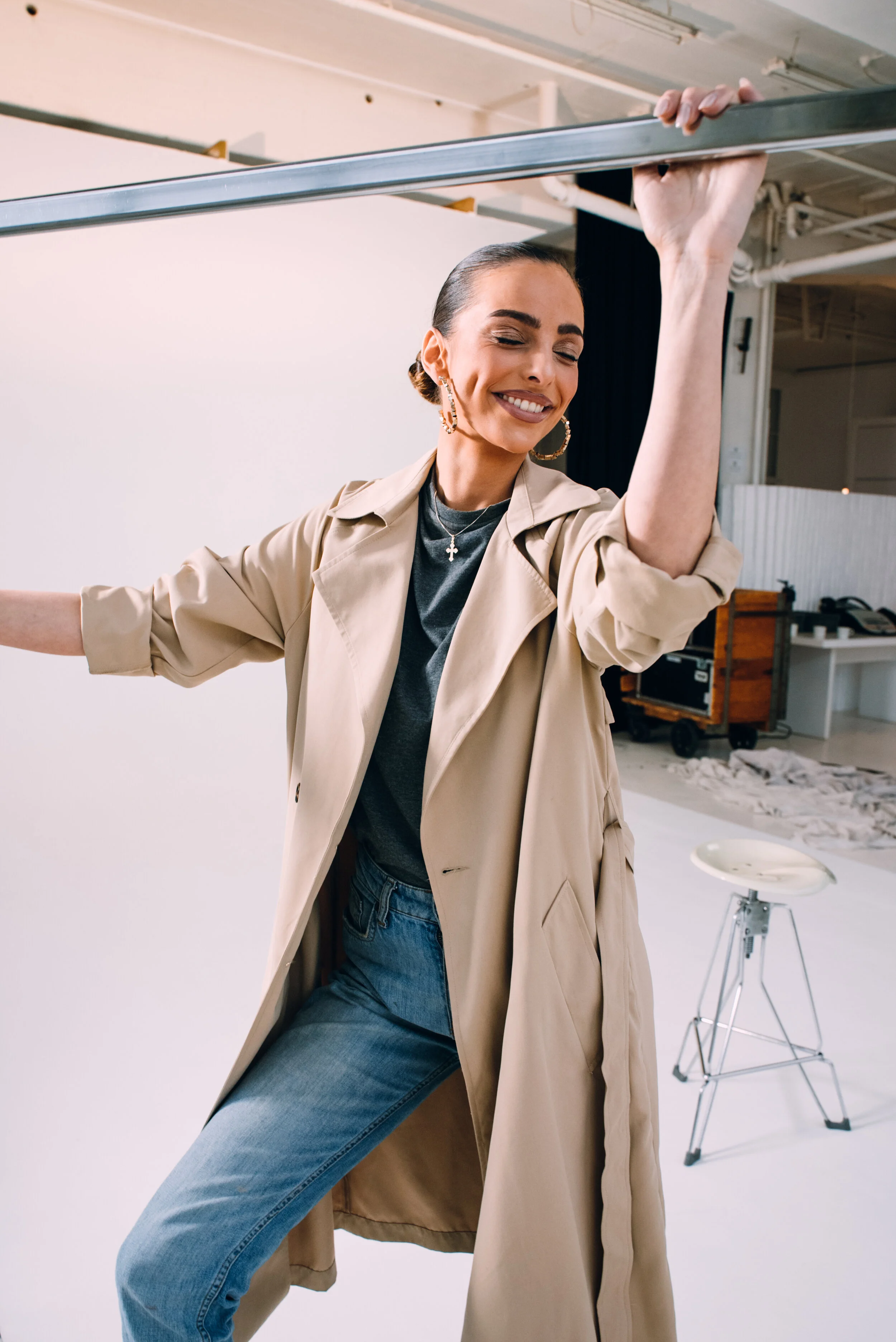 A woman in a beige trench coat and jeans holding a metal bar, likely in a photo studio or art space, smiling with eyes closed.