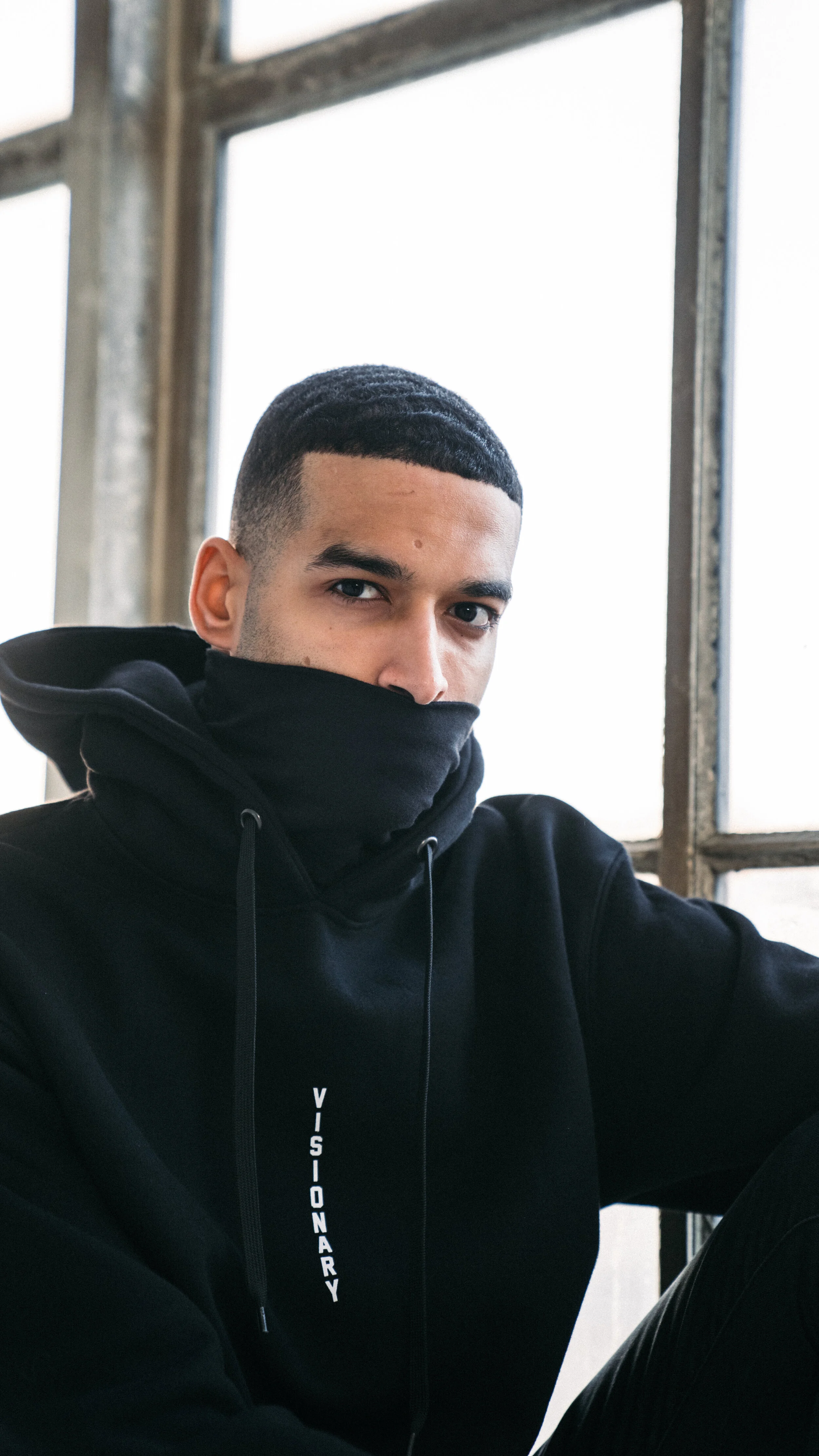 A young man wearing a black hoodie with the word 'VISAIONARY' on it, and a matching black face mask covering his mouth and nose, sitting near large windows with a metal frame and bright light coming through.