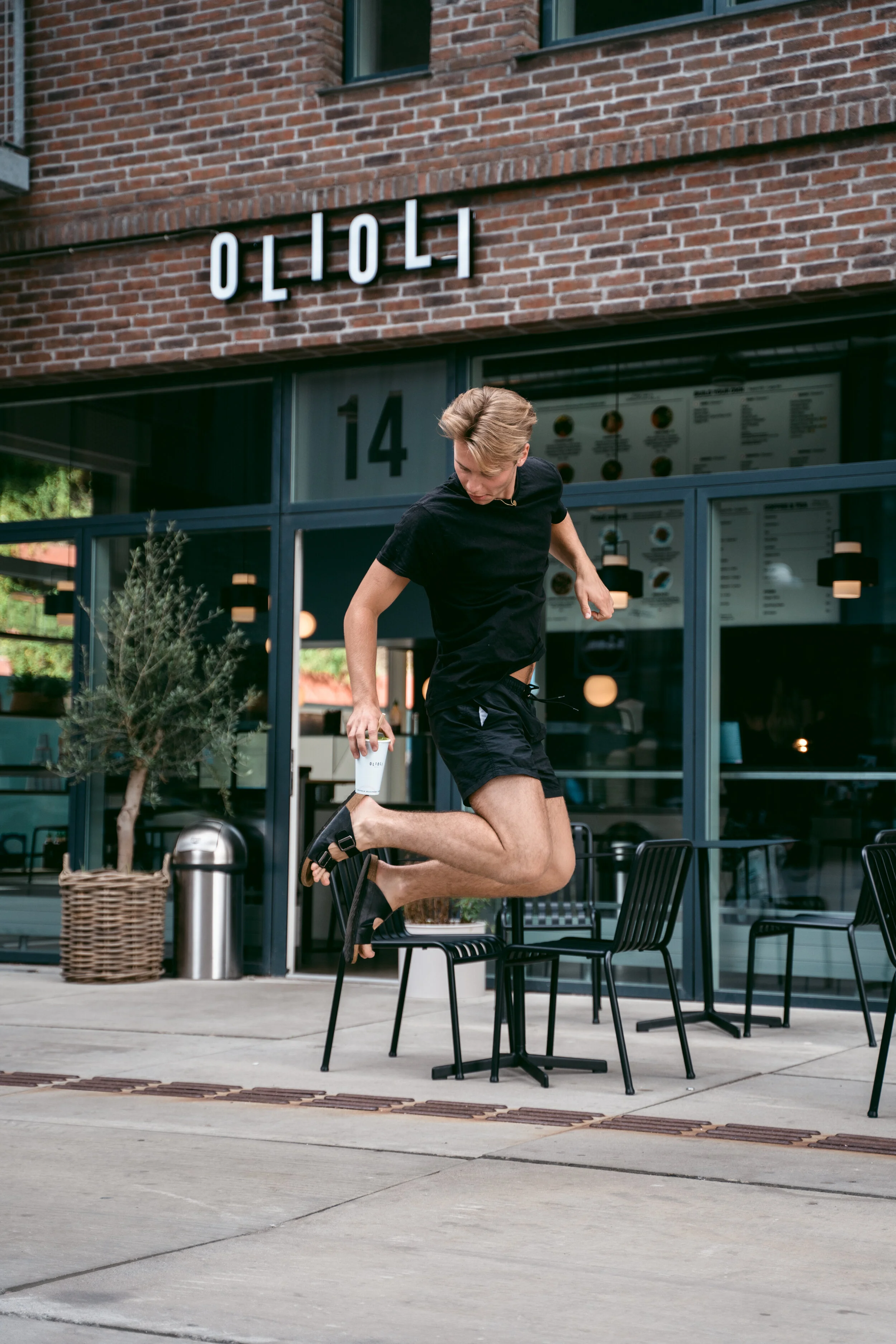 A young man jumping onto a chair outside a modern cafe or restaurant.