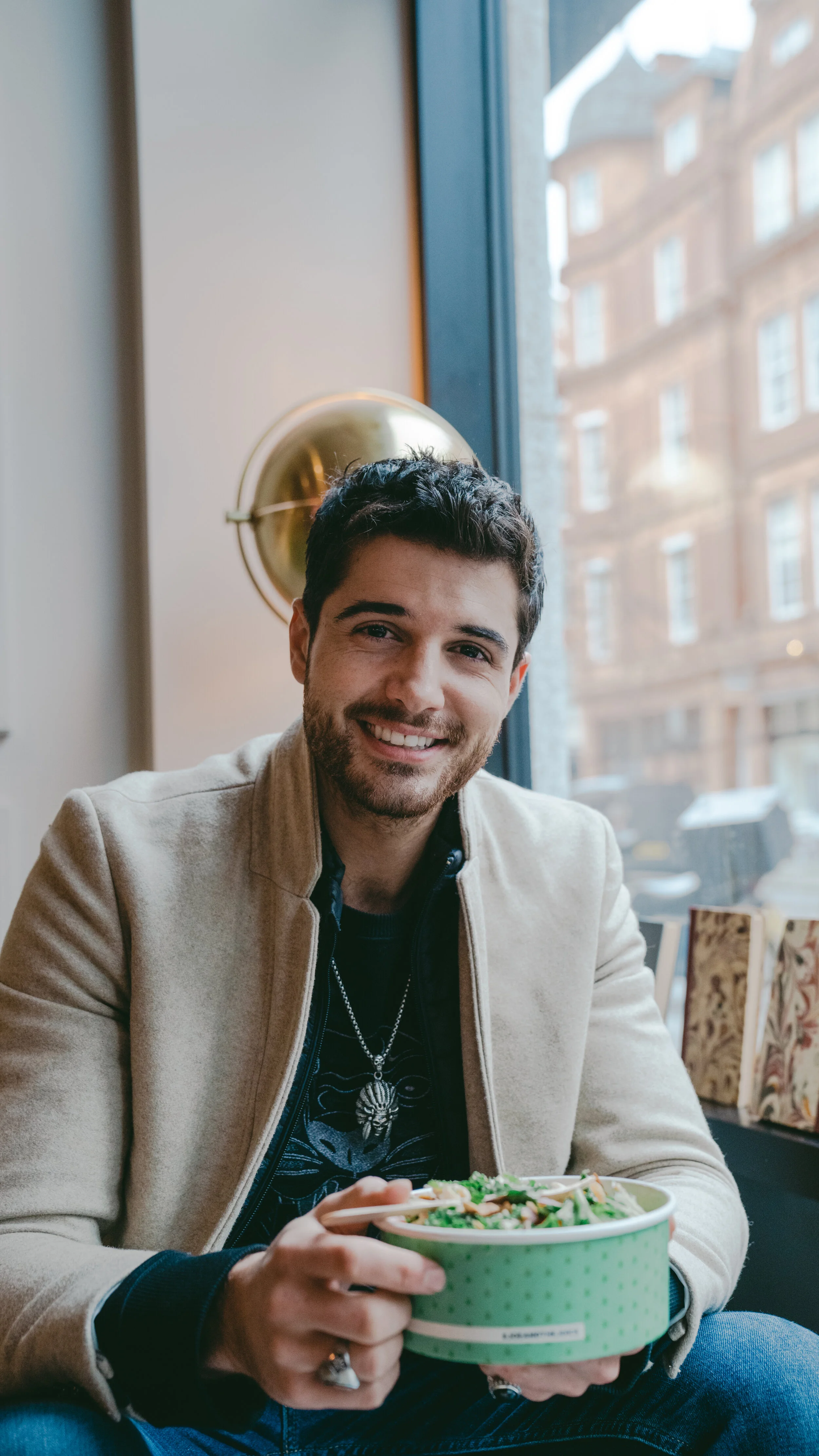 A man smiling and holding a bowl of salad while sitting indoors near a window with a city view.