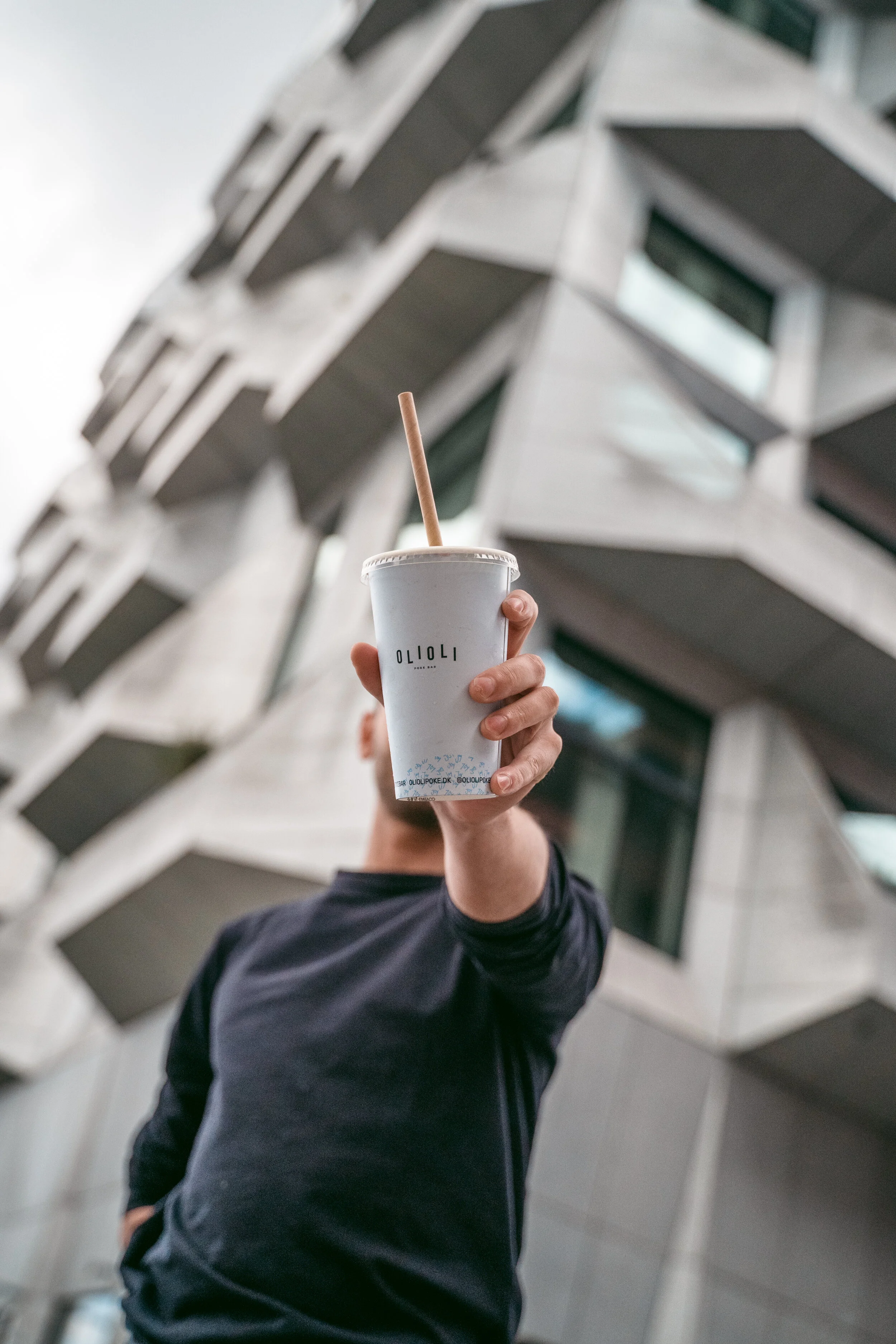 Person holding a white disposable cup with a straw in front of a modern building with angular architecture.