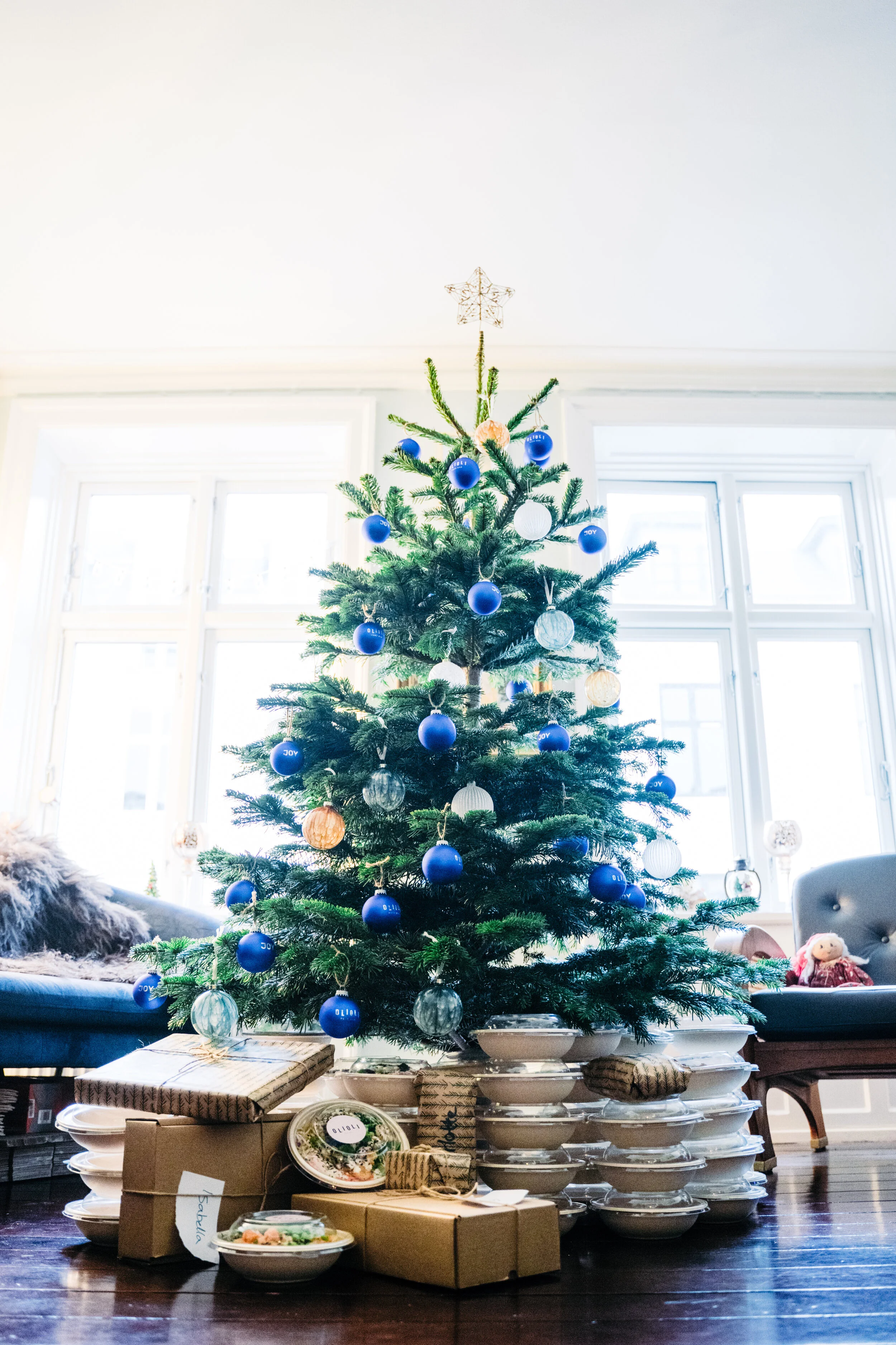 A decorated Christmas tree with blue and silver ornaments, topped with a star, in a room with large windows and presents underneath.