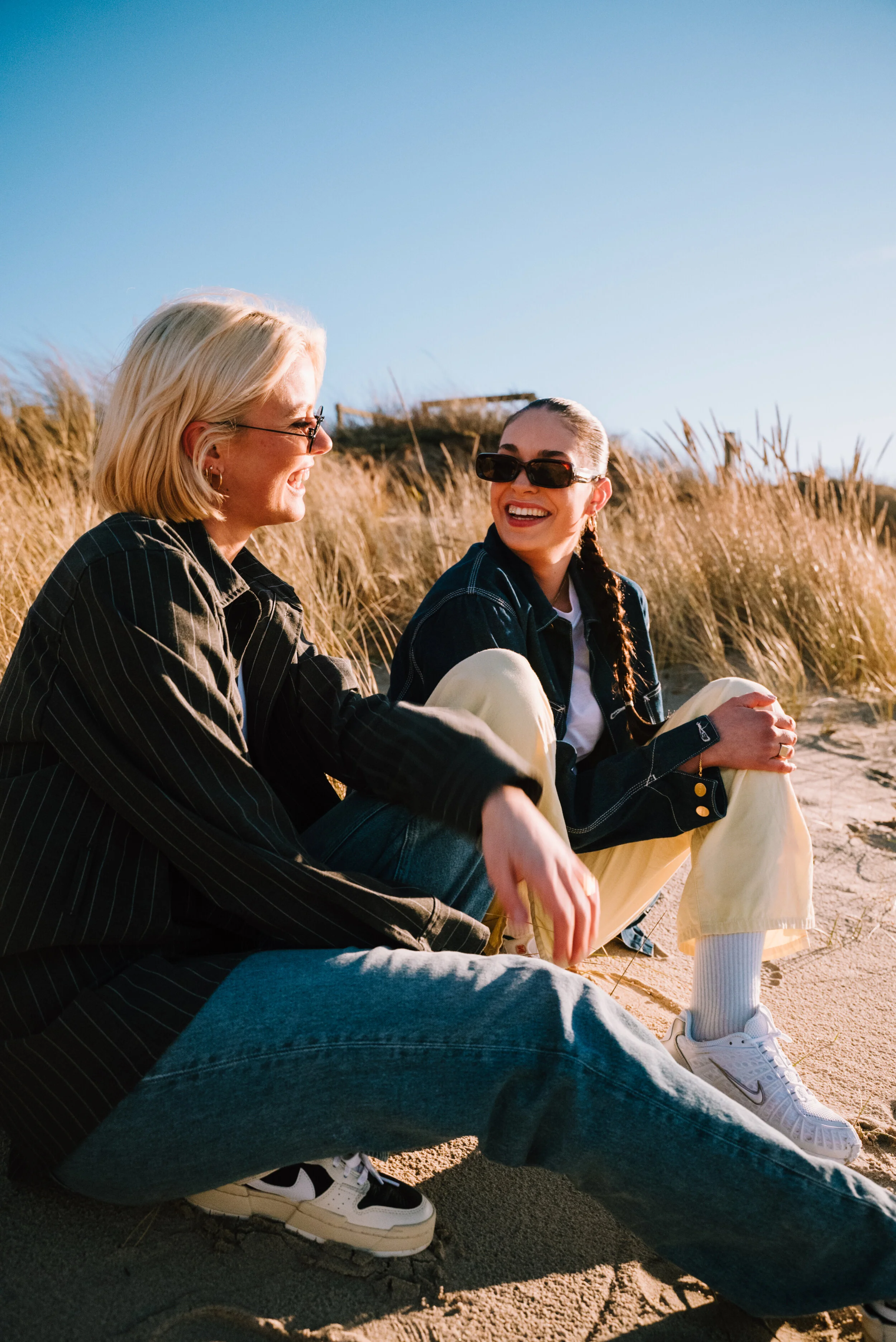 Two women sitting on a sandy beach, laughing and enjoying each other's company during sunset, with tall grass behind them.
