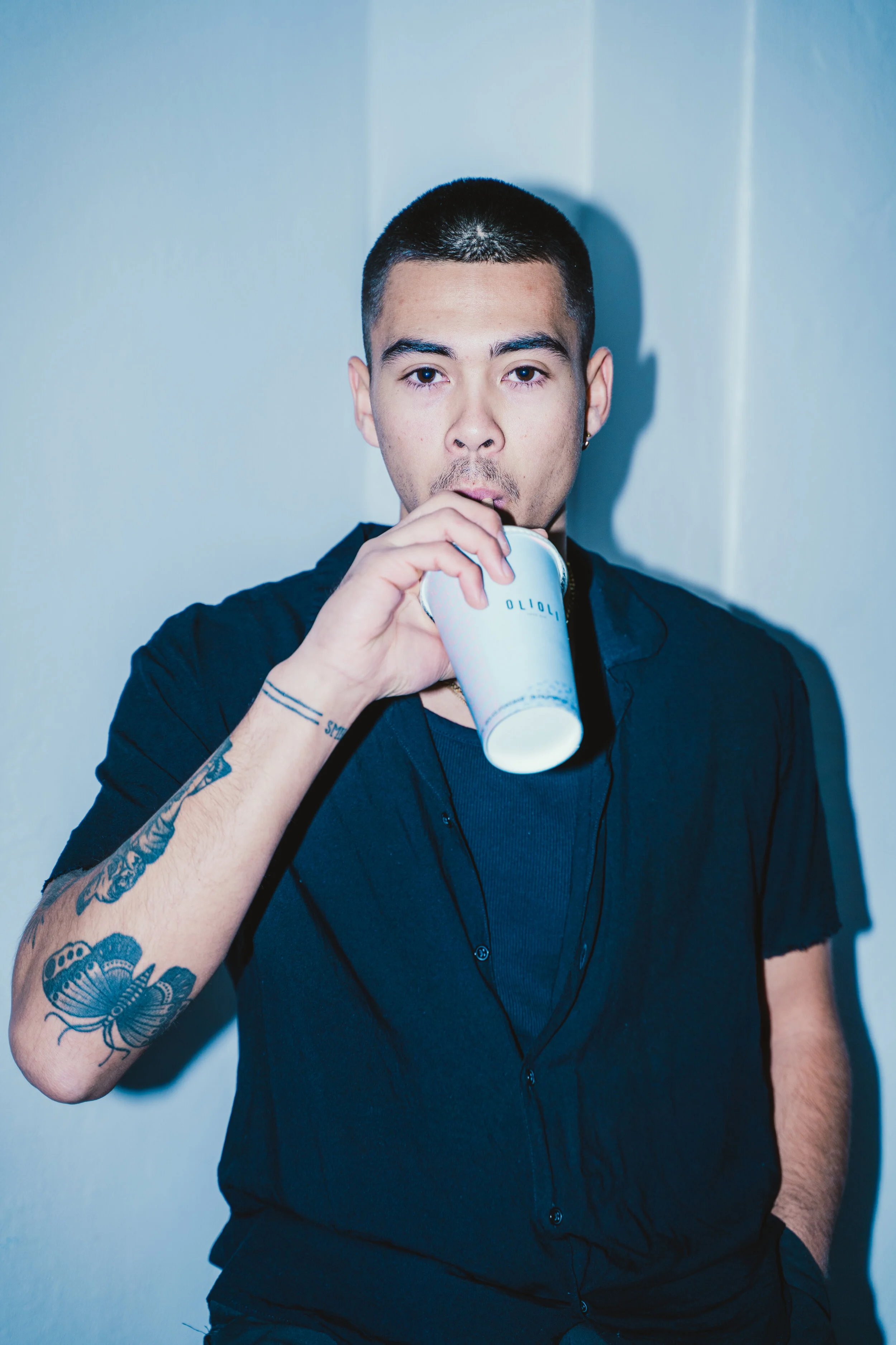 A young man with short hair, tattoos on his arm, wears a dark shirt, drinks from a white cup, standing against a light-colored wall.