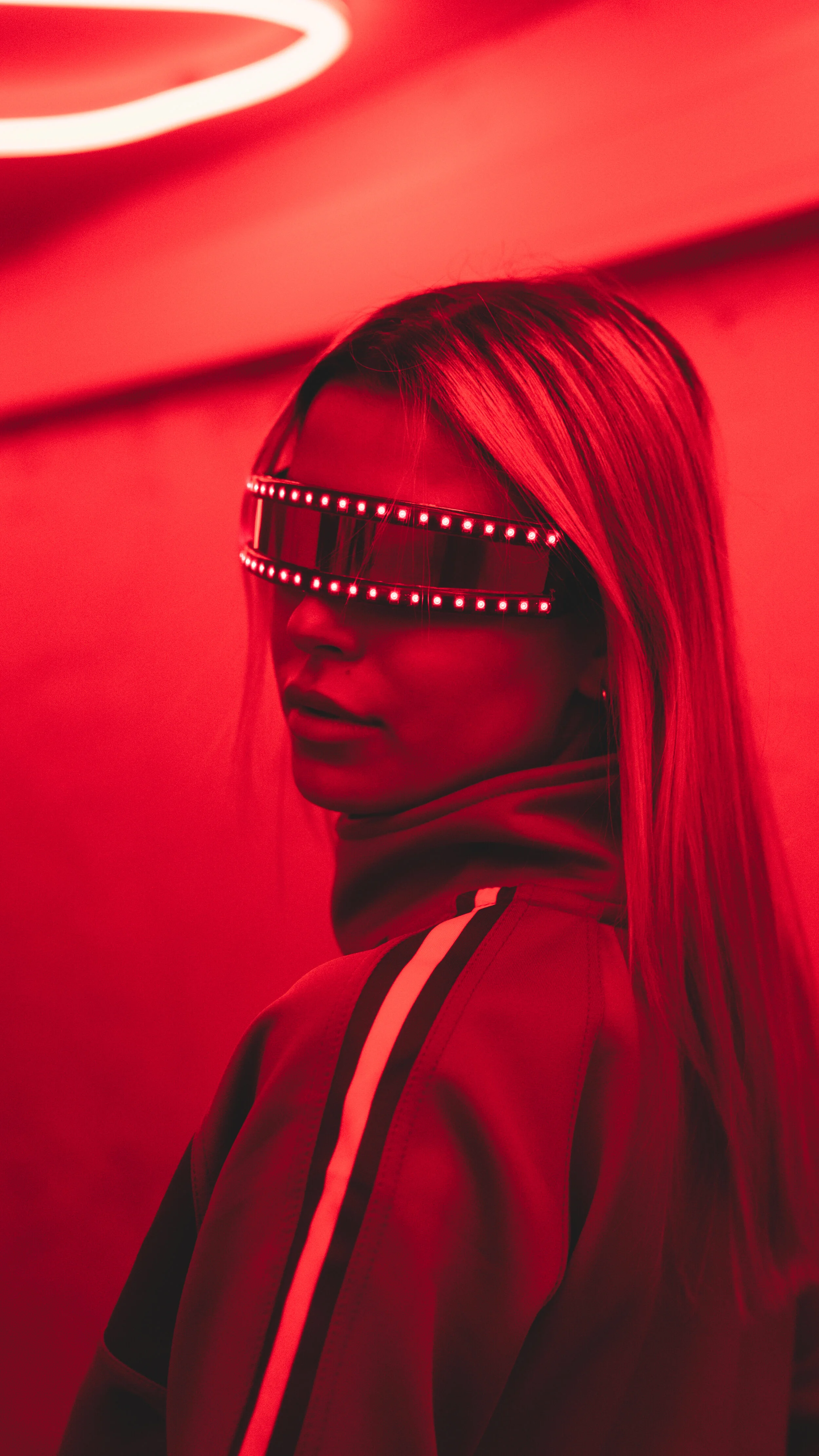 A woman with long hair wearing LED sunglasses that cover her eyes, in a red-lit setting, with a red and white striped sports jacket.