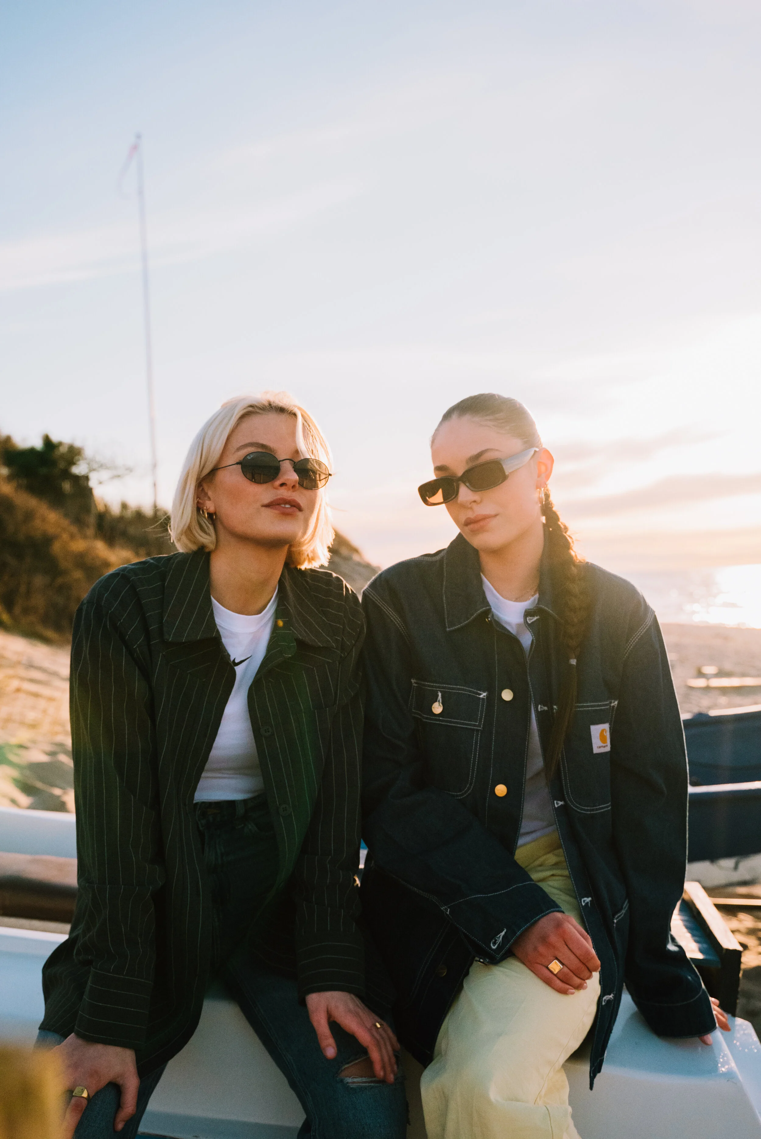 Two women with sunglasses sitting on a boat at sunset near the beach, wearing casual jackets and white shirts.