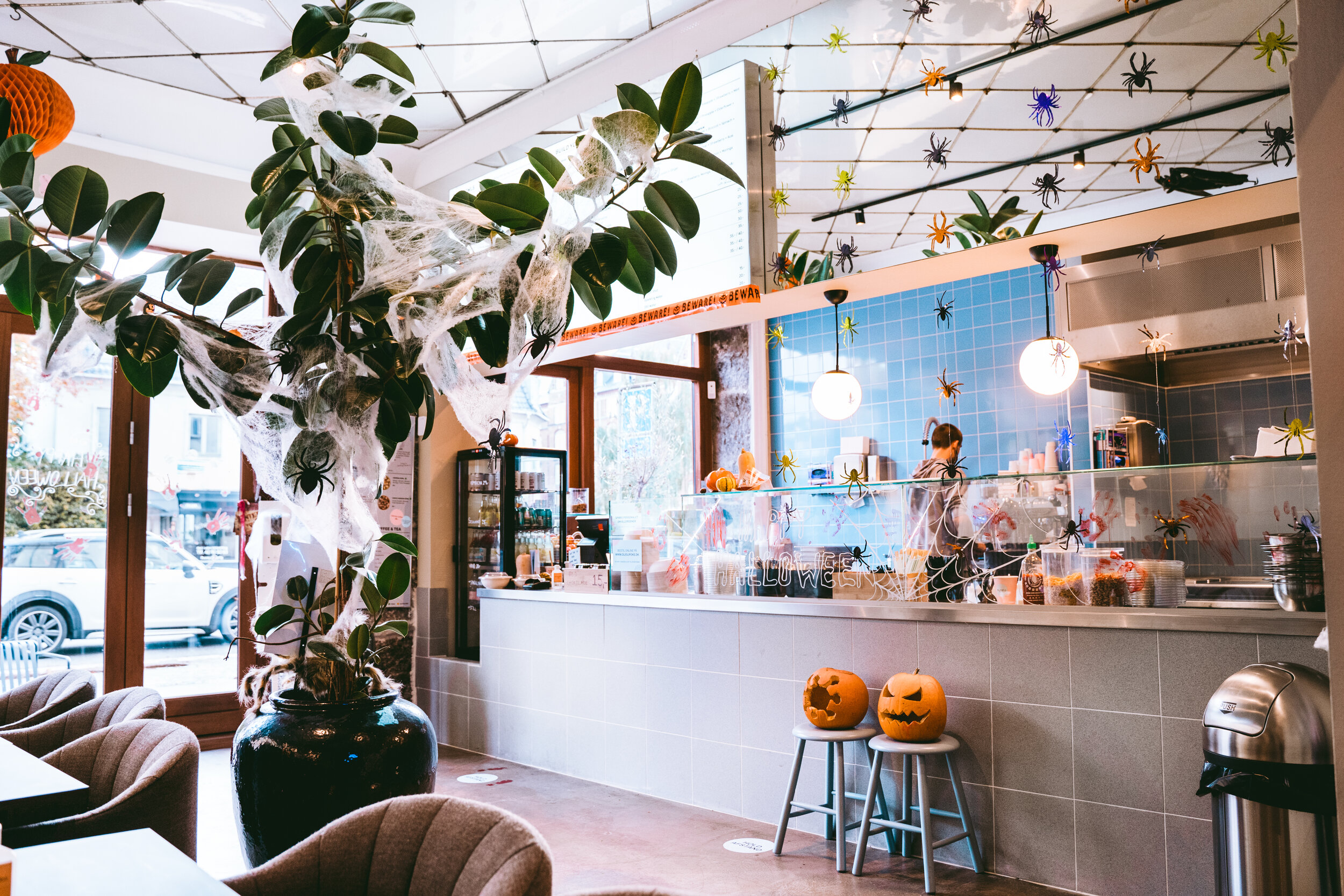 Cafe decorated for Halloween with spiderwebs, fake spiders, and carved pumpkins, featuring a counter area with a person working, Halloween decorations hanging from the ceiling, and chairs near large windows
