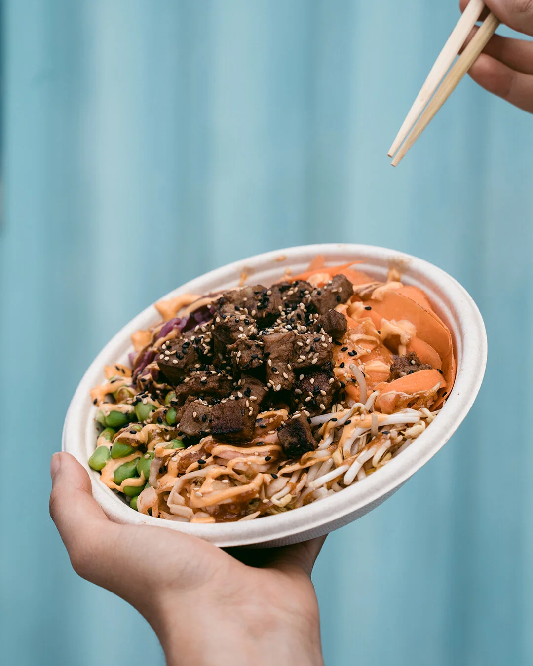 A hand holding a white bowl of Asian noodle salad topped with meat, sesame seeds, and vegetables, with chopsticks in the other hand.