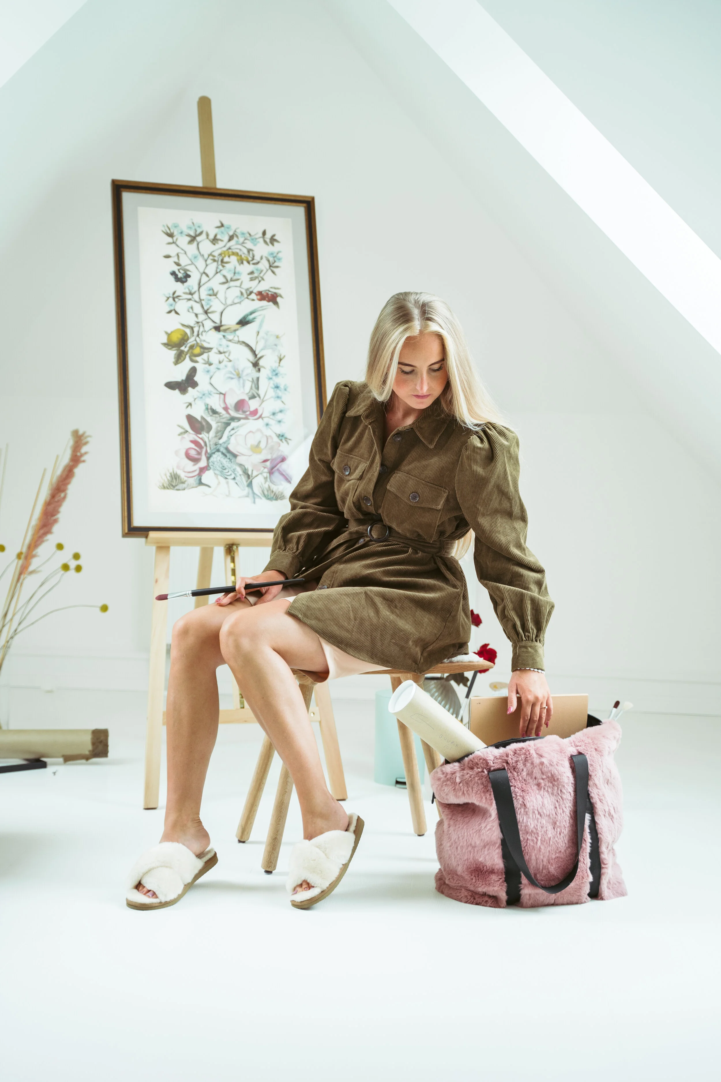 A woman in a brown dress with puffed sleeves sitting on a wooden stool in an art studio. She holds a paintbrush in one hand and looks down. Behind her is an easel with a large framed botanical painting, and a pink fluffy tote bag with art supplies on