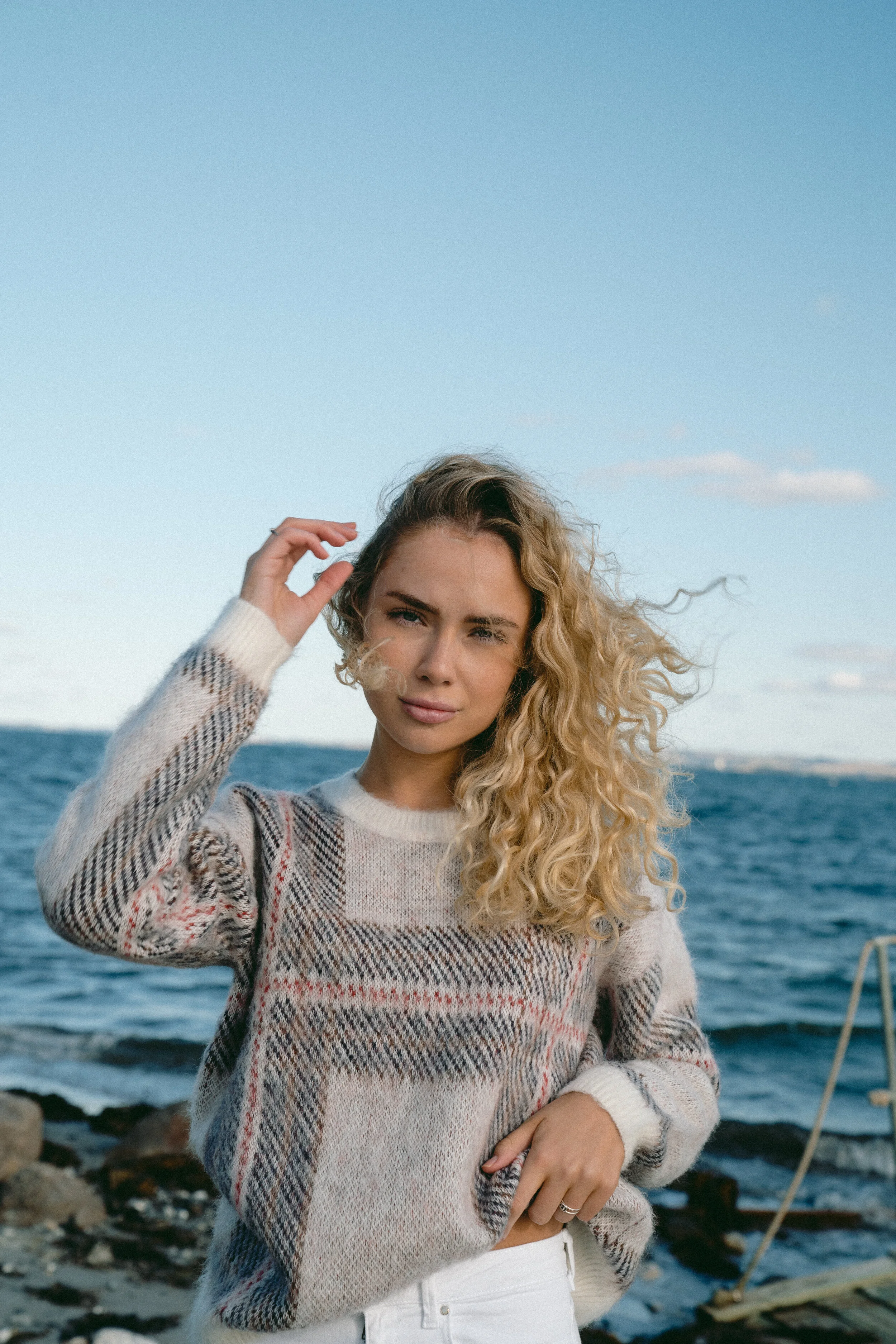 Young woman with curly blonde hair standing on a rocky shoreline near the ocean, wearing a patterned sweater and white pants, with wind blowing her hair.