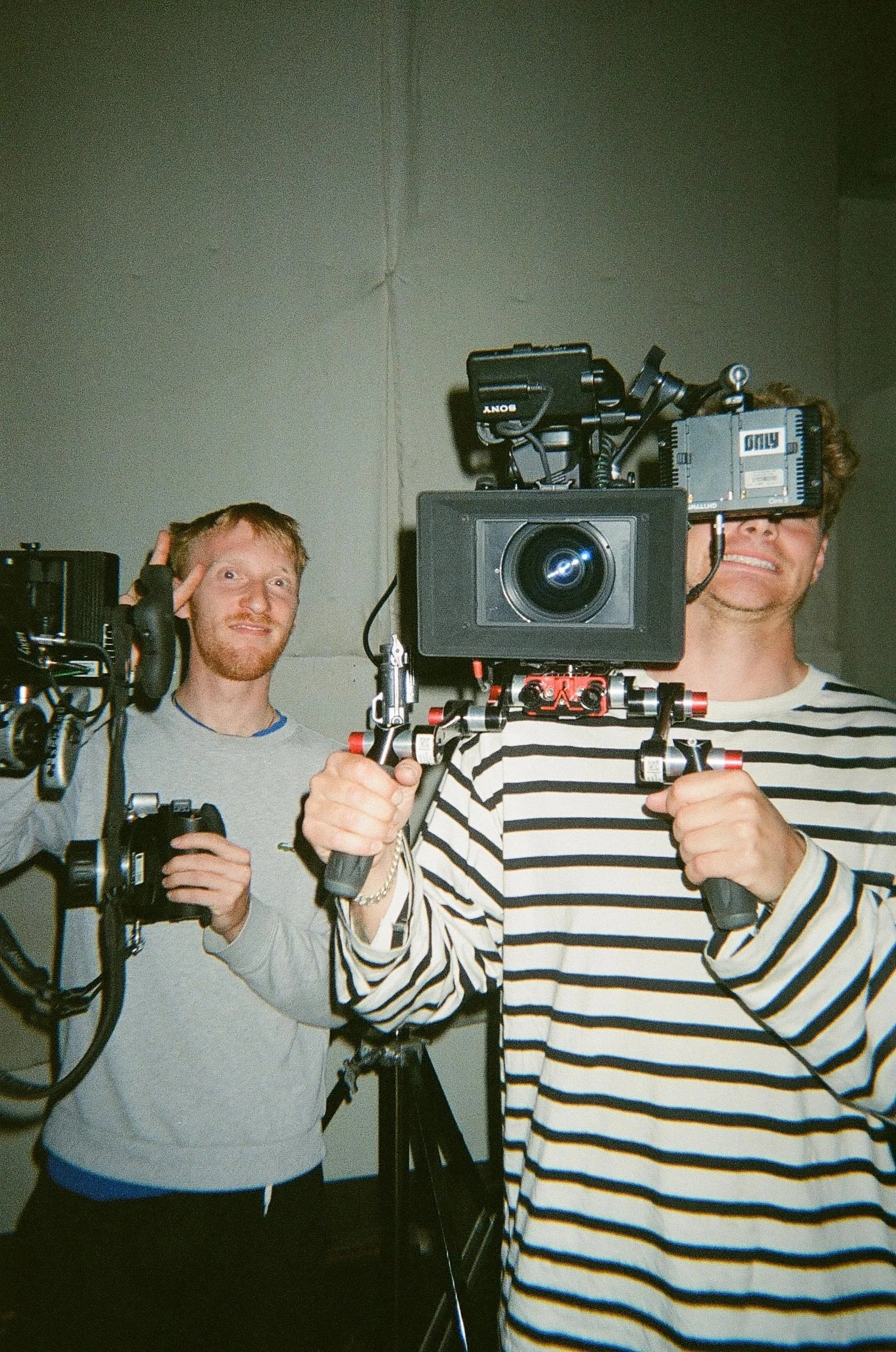 Two men operating professional video recording equipment in a room with gray walls, one smiling and the other making a peace sign.