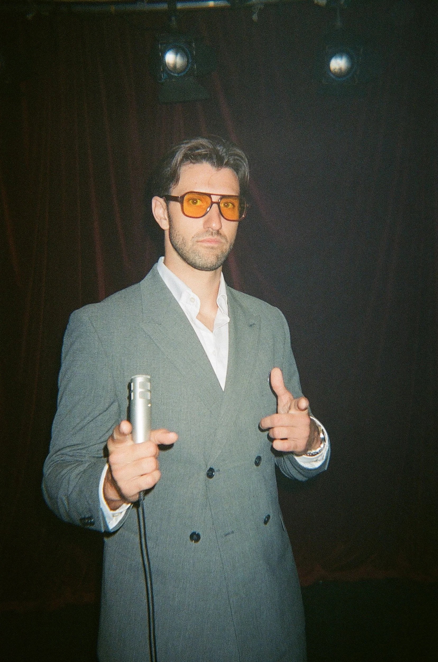 A man in a gray double-breasted suit and white shirt with a few buttons open, wearing orange-tinted sunglasses, pointing with both hands, holding a microphone in his right hand, standing in front of dark curtains with stage lights above.