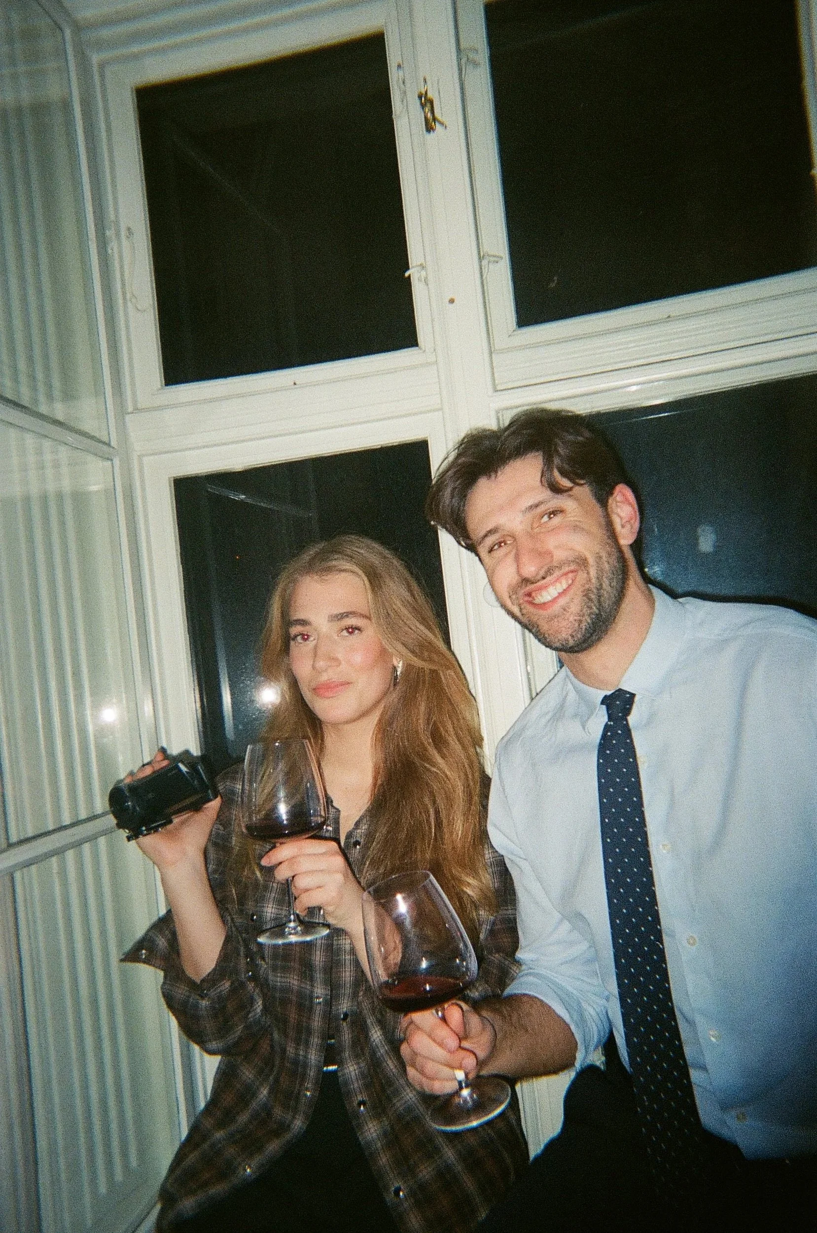 Young woman and man smiling, holding glasses of red wine, in a room with large window panes.
