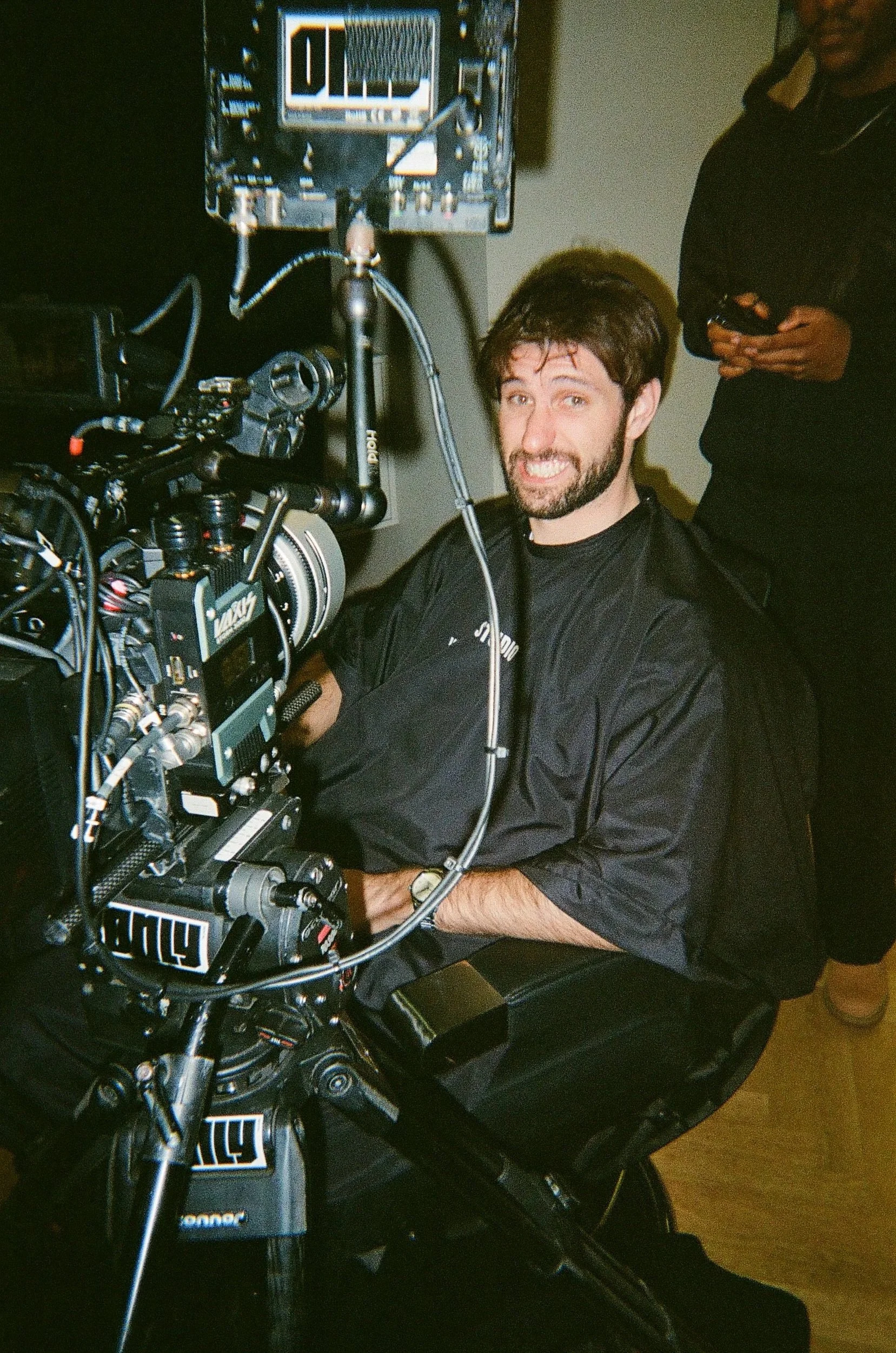 A smiling man with a beard sitting in front of a professional film camera, with a crew member standing behind him using a phone.