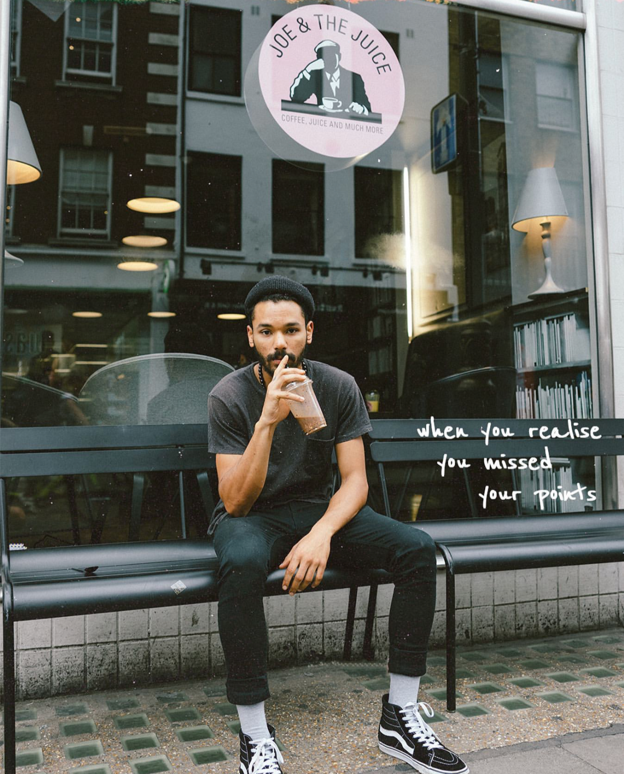 A young man with a beard and wearing a black beanie, black t-shirt, black pants, white socks, and Vans sneakers, sitting on a black bench outside a cafe, drinking a smoothie. The cafe window displays the logo 'Joe & The Juice' and a quote that says '