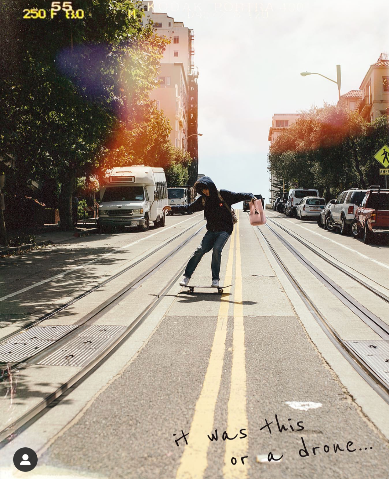 A person skateboarding on double yellow lines on a city street with parked cars and trees, sunny day with lens flare, handwritten note at the bottom saying 'it was this or a drone...'