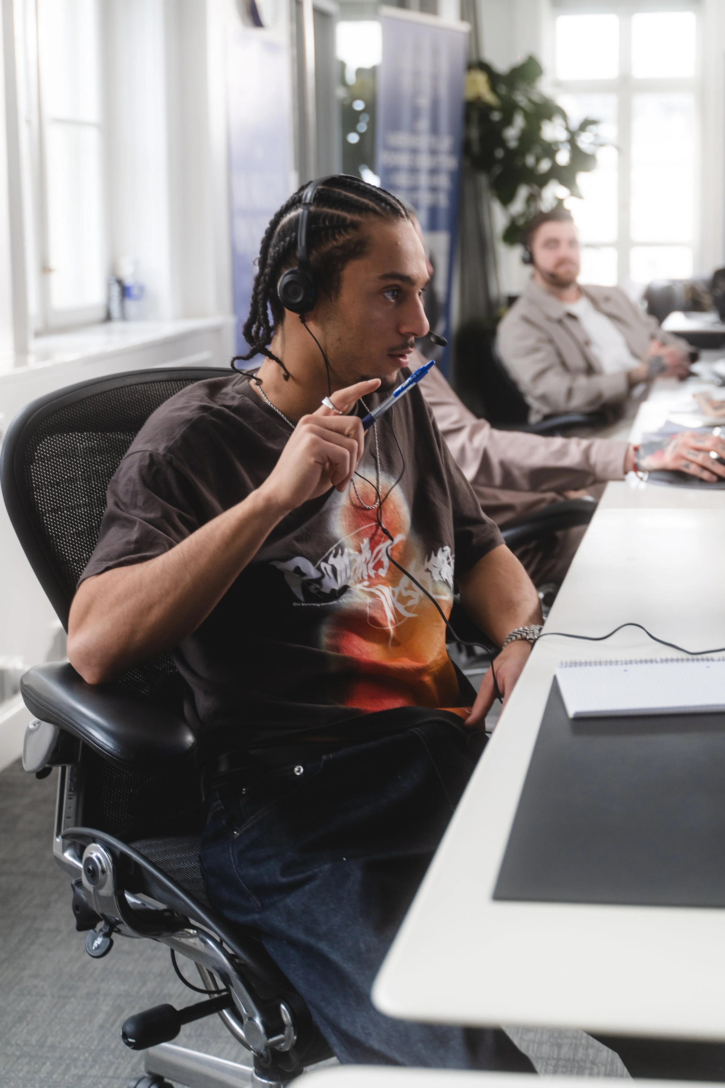 Young man with braids wearing a headset, holding a pen, and sitting at a conference table with laptops and notebooks, in an office with large windows.
